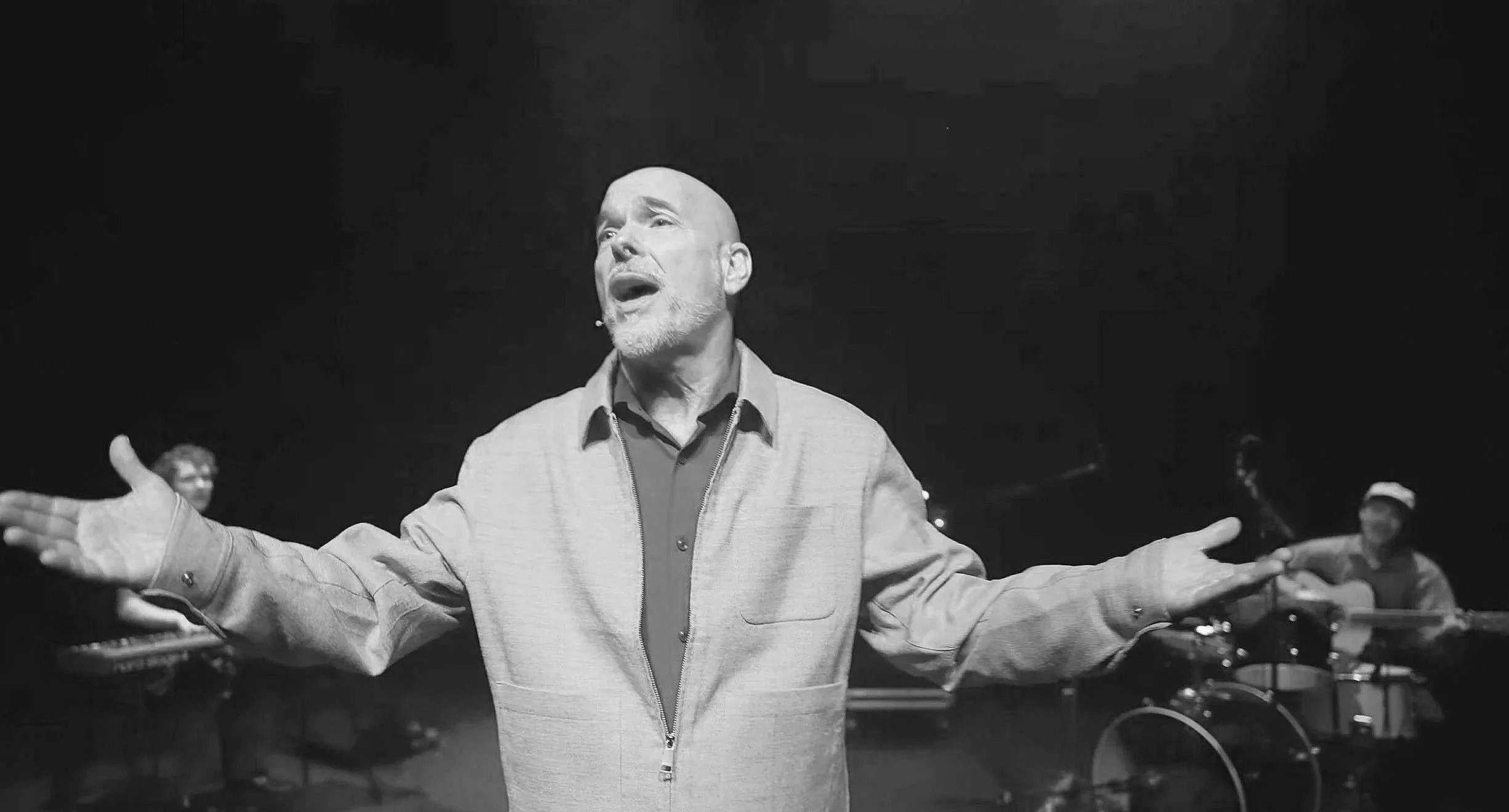 Een man met een grijze baard zingt of spreekt met zijn arm uitgestoken, terwijl er muzikanten met instrumenten op de achtergrond staan in een donkere omgeving.