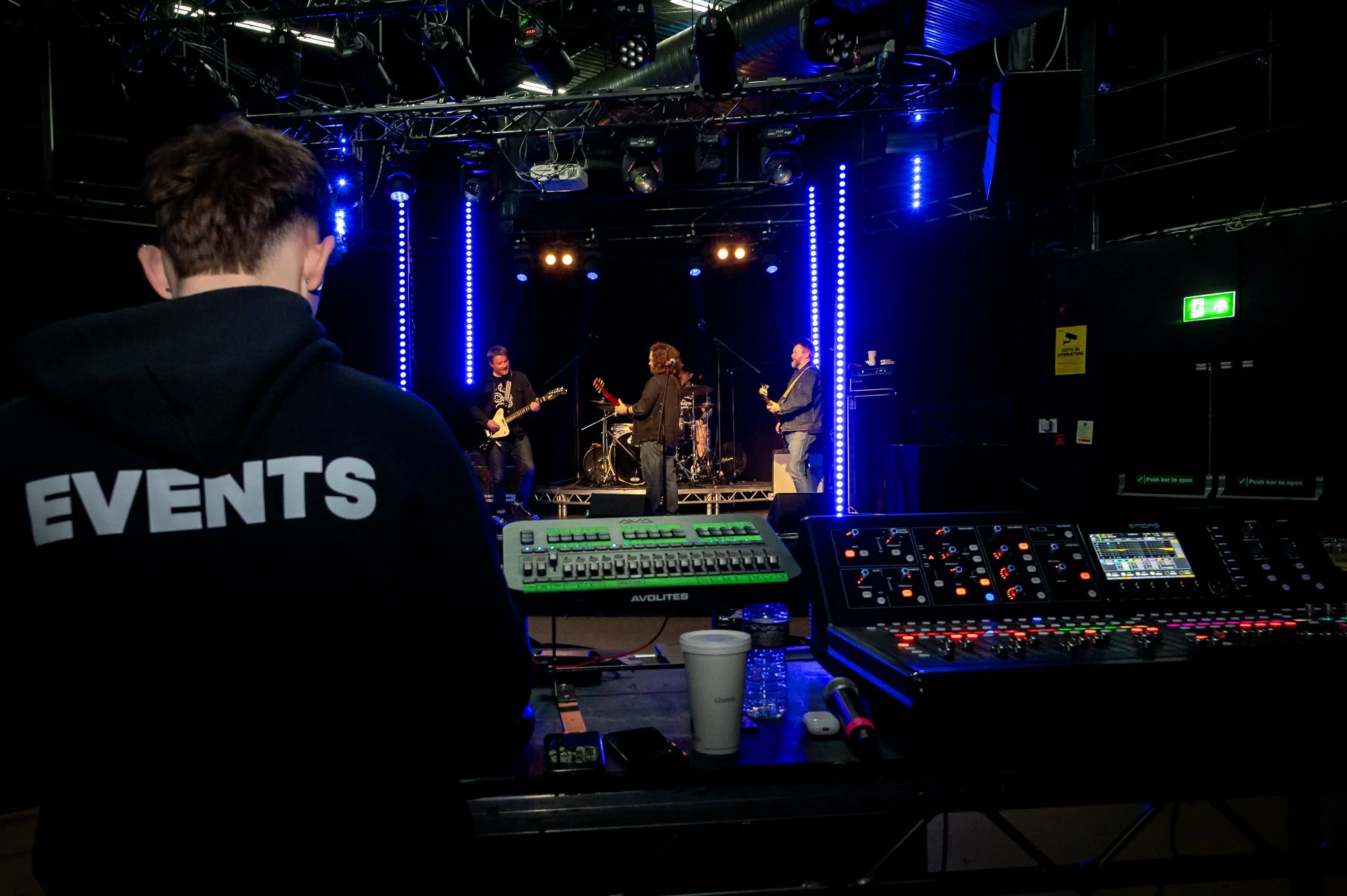 A crew member wearing a hoodie with 'EVENTS' on the back oversees a live band performance on stage, with music equipment and controls in the foreground, blue stage lights, and a dark venue background.