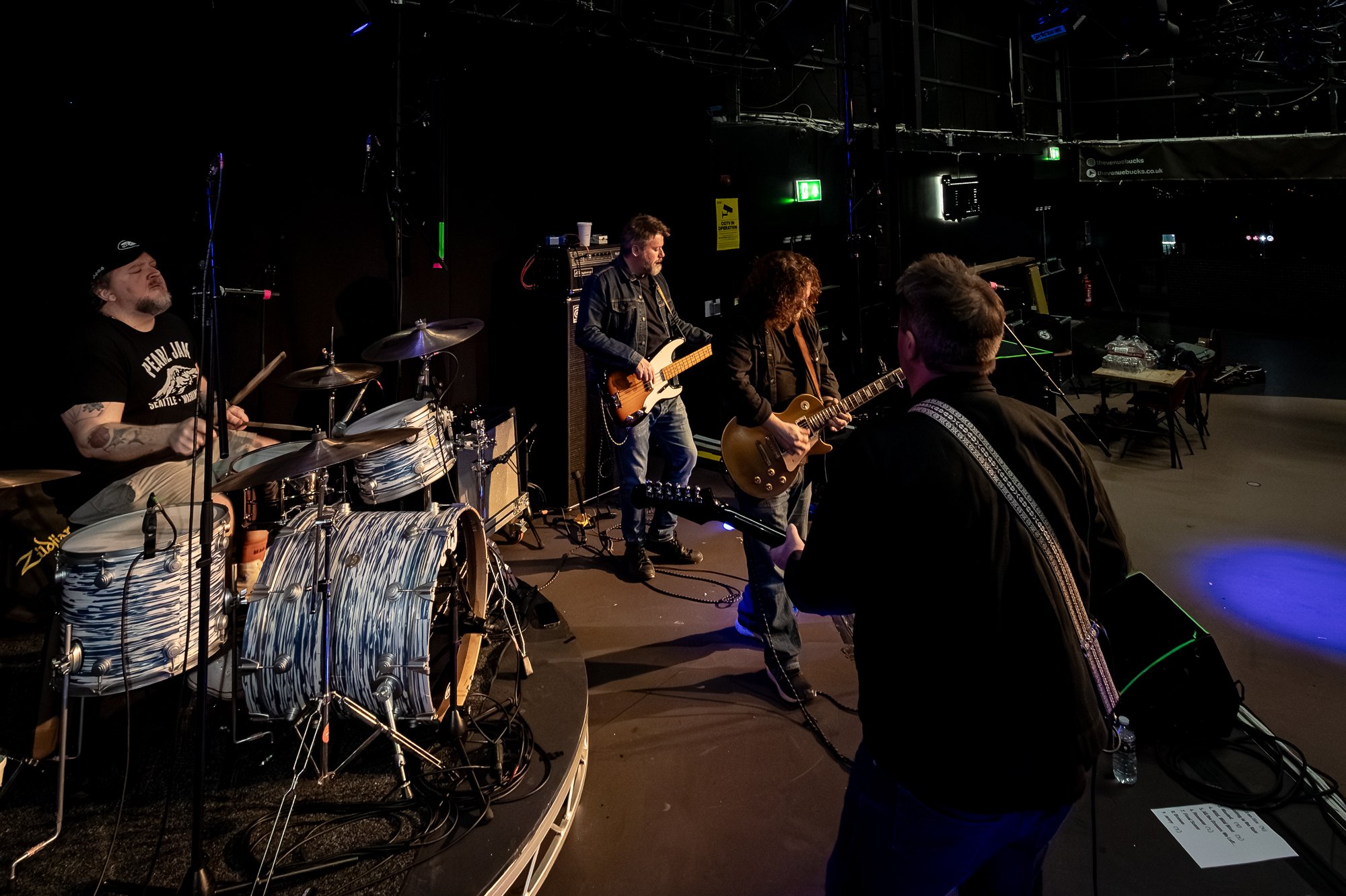 A band performing on stage, featuring a drummer, two guitarists, and a person in the foreground with a guitar, in a dimly lit venue.