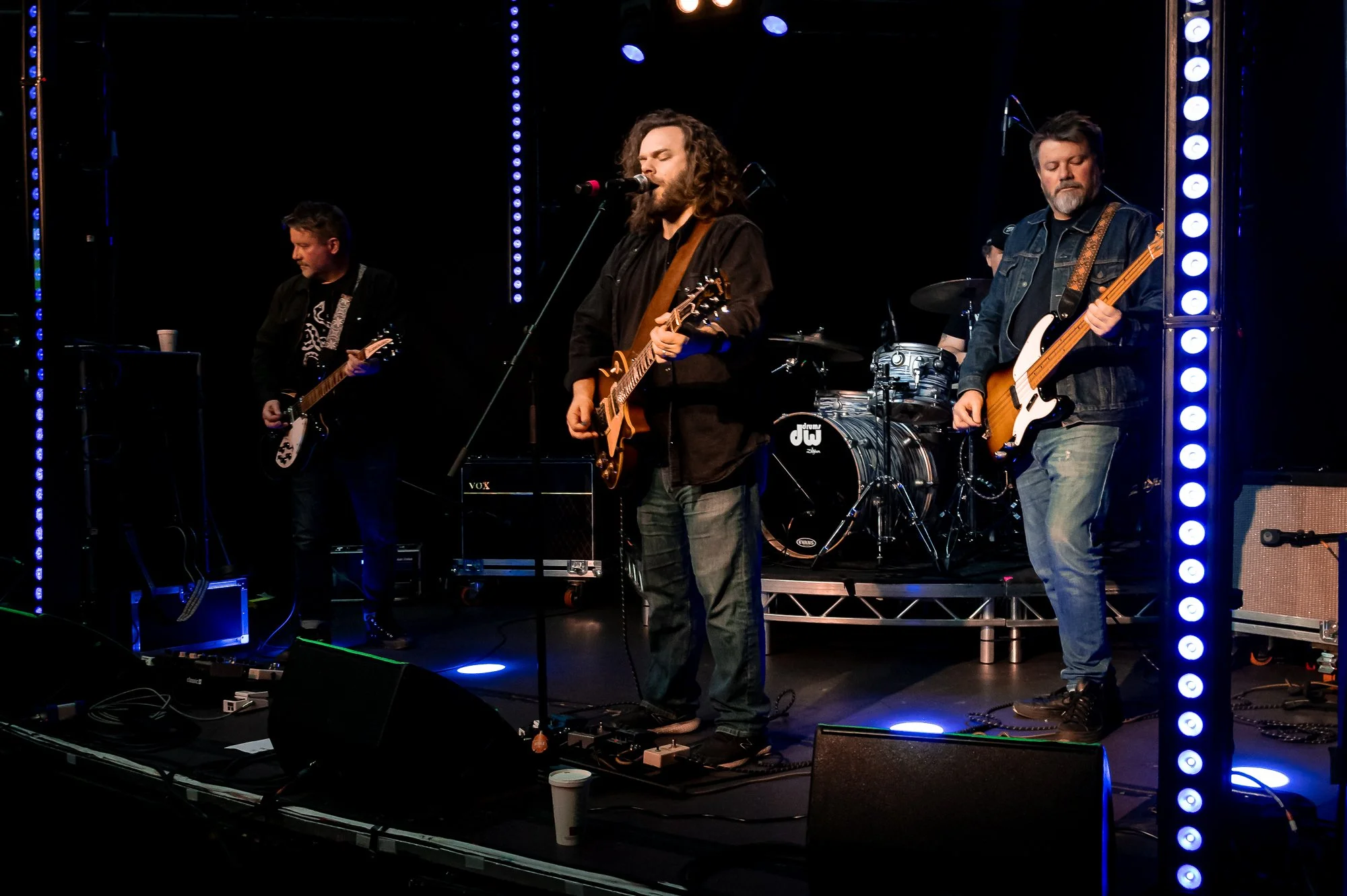 A band of four musicians performing on a stage with blue lighting, including a guitarist, a lead singer with a guitar, another guitarist, and a bassist. There is a drum set in the background, and various music equipment on stage.