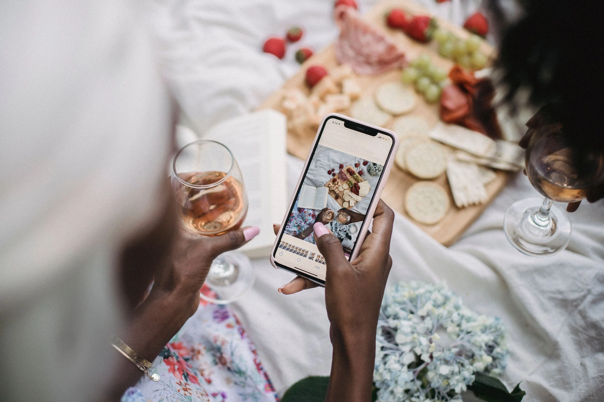 Person taking a picture of a cheeseboard with their phone while holding a glass of wine