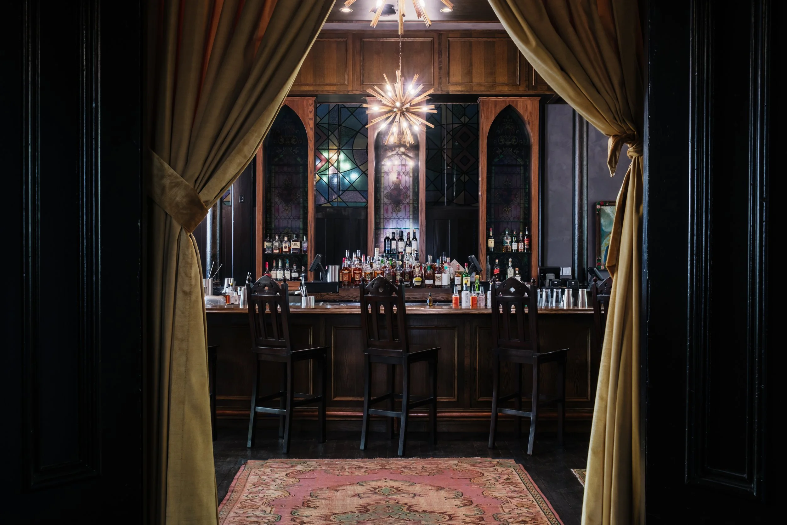 The bar at The Last Refuge, framed by velvet curtains and warm wood tones, showcasing an intimate bourbon bar setting inside a restored church in Louisville, Kentucky.