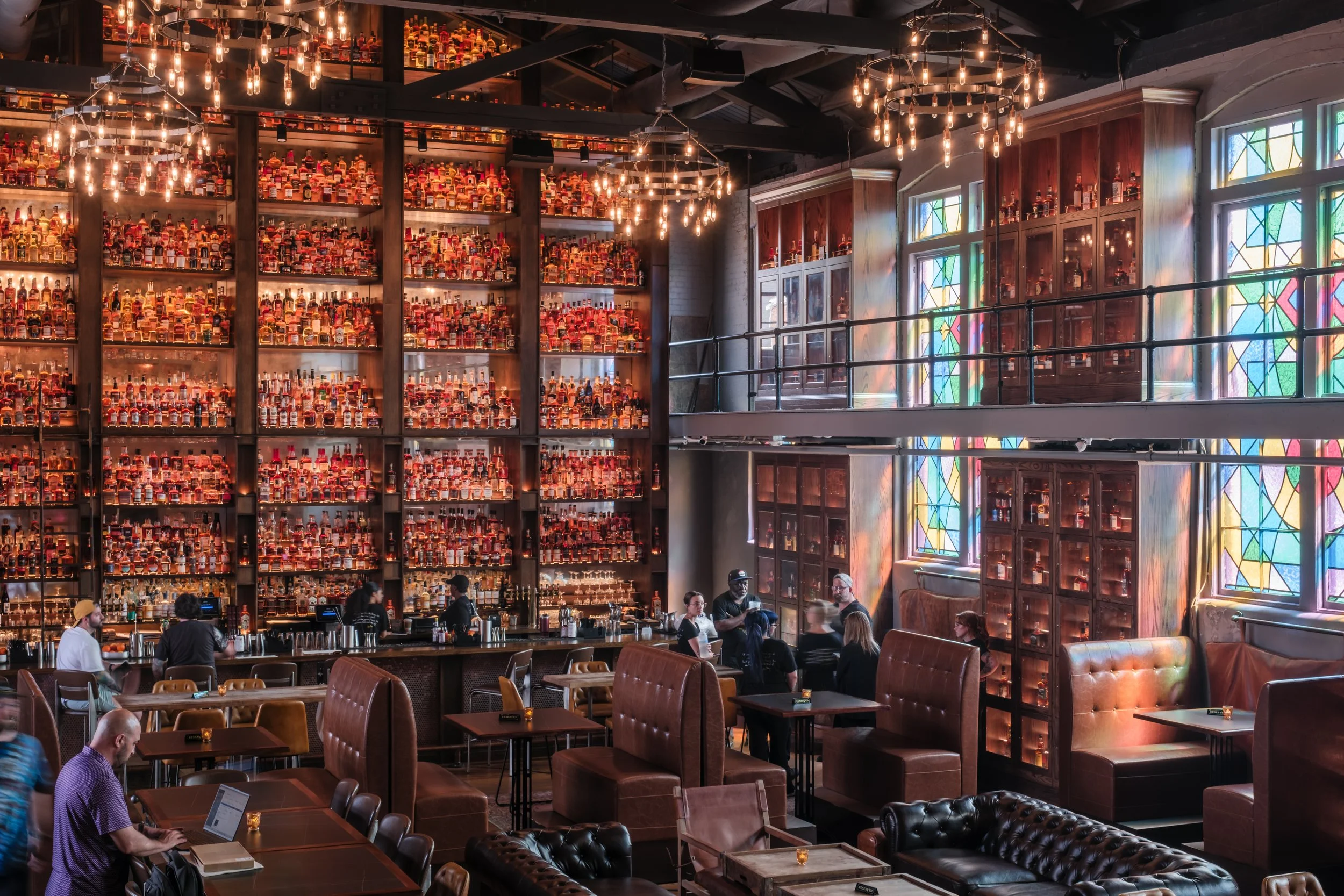 Guests enjoying cocktails beneath towering bourbon shelves at The Last Refuge in Louisville, highlighting the venue’s energetic yet refined atmosphere during peak evening hours.
