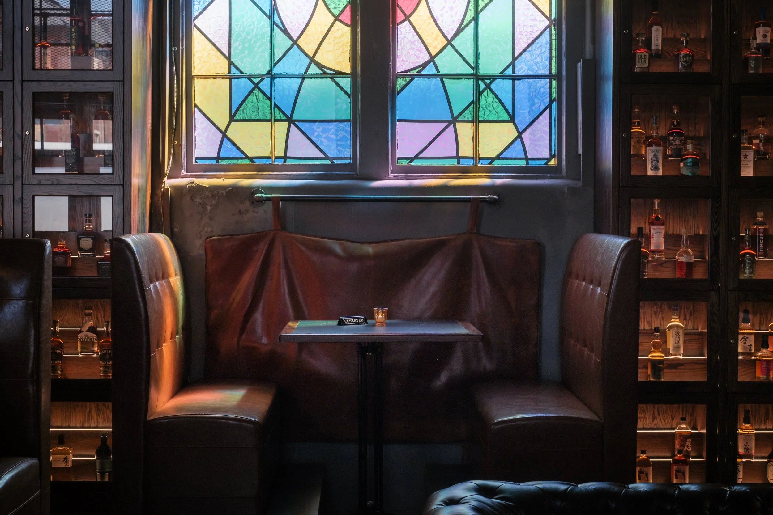 A cozy booth beneath stained-glass windows at The Last Refuge, blending historic church architecture with an intimate bourbon and cocktail lounge atmosphere in Louisville.
