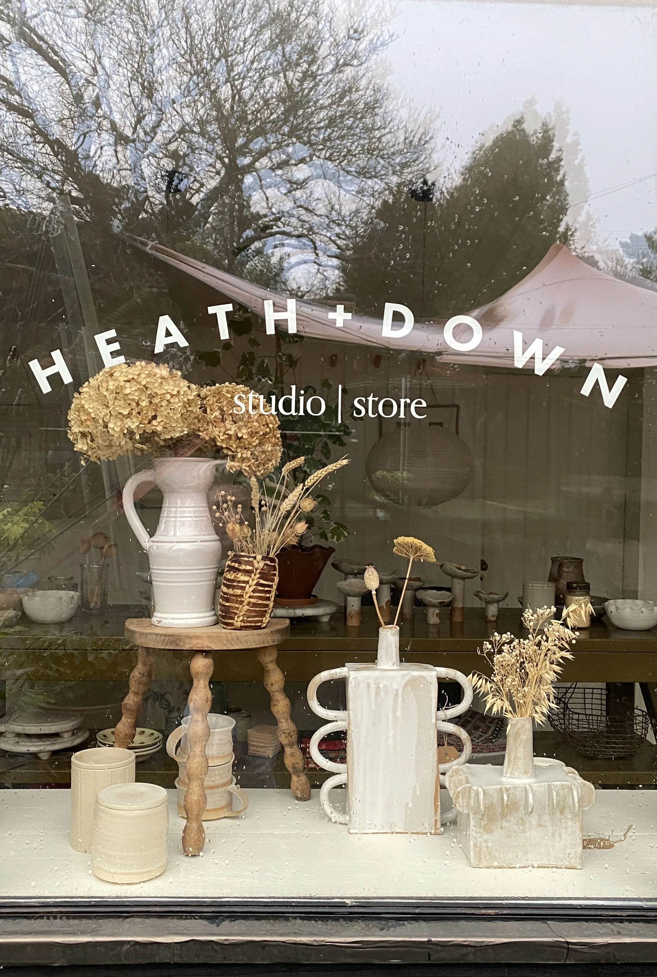 A store window display with dried flowers in vases, a small wooden table, and ceramic bowls. The store is called 'Heath + Down' and offers both studio and retail services. East Sussex.