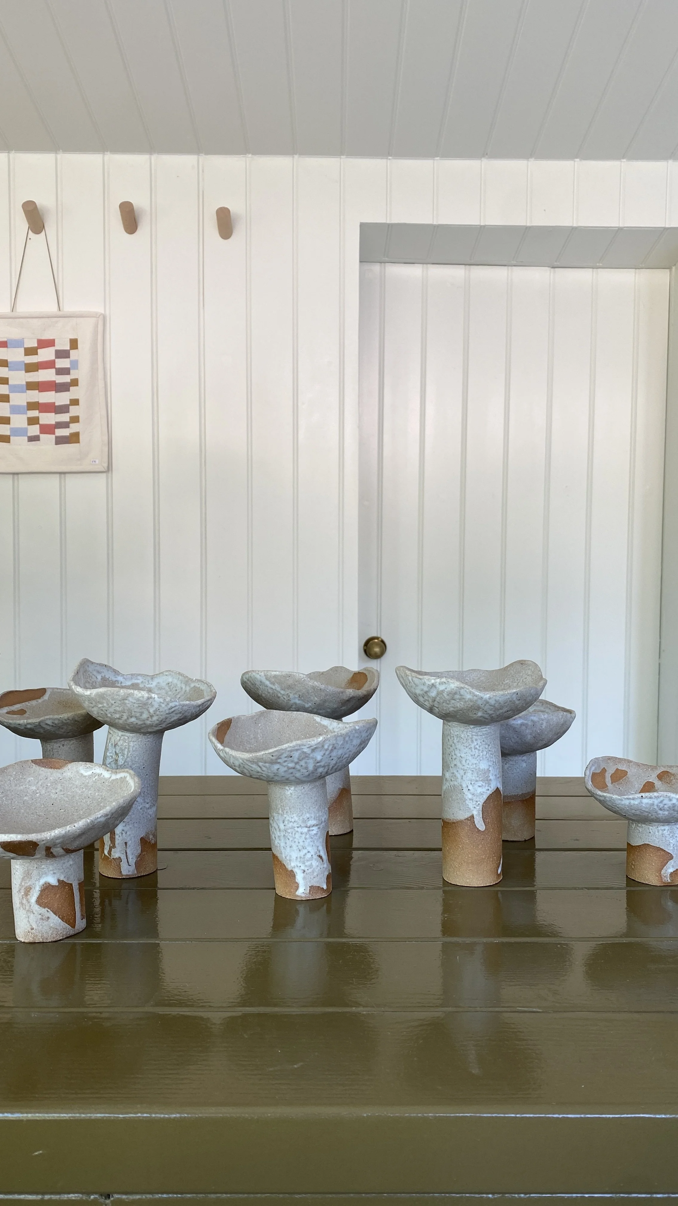 Collection of ceramic mushrooms on a wooden table in a room with white paneled walls and a white door. Handmade. Homemade. Sussex artist.