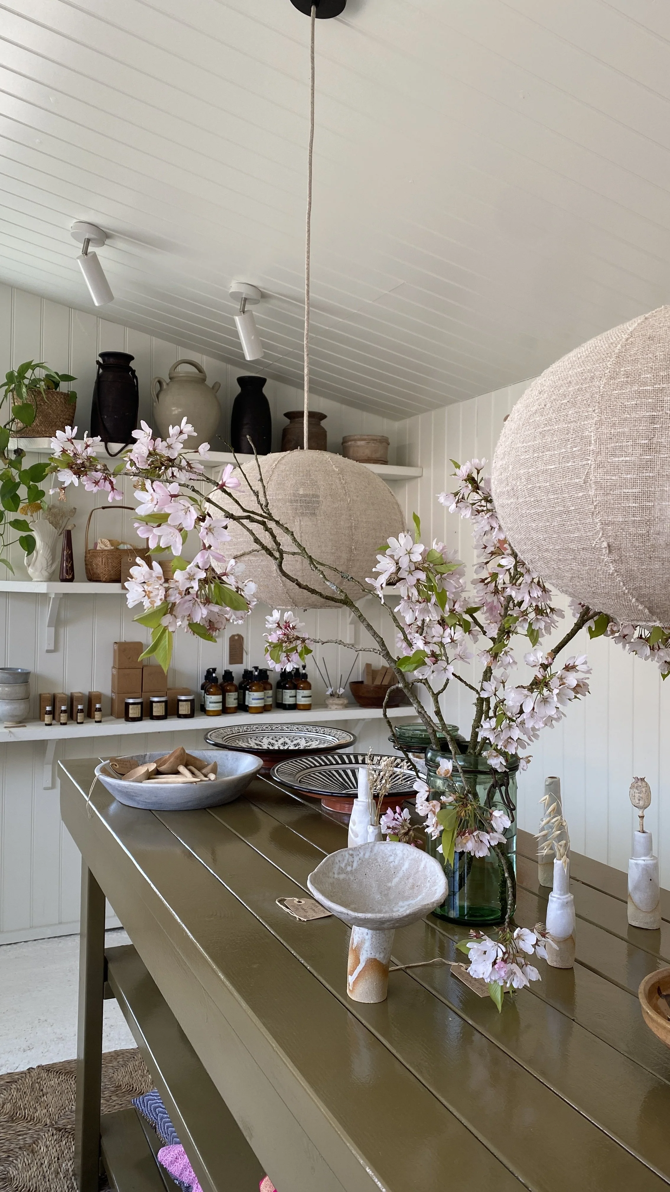 Interior of a room with a wooden dining table or countertop decorated with ceramic and glass vases, bowls, and candles. Hanging linen lampshades, shelves with pottery and bottles. Interior design East Sussex.