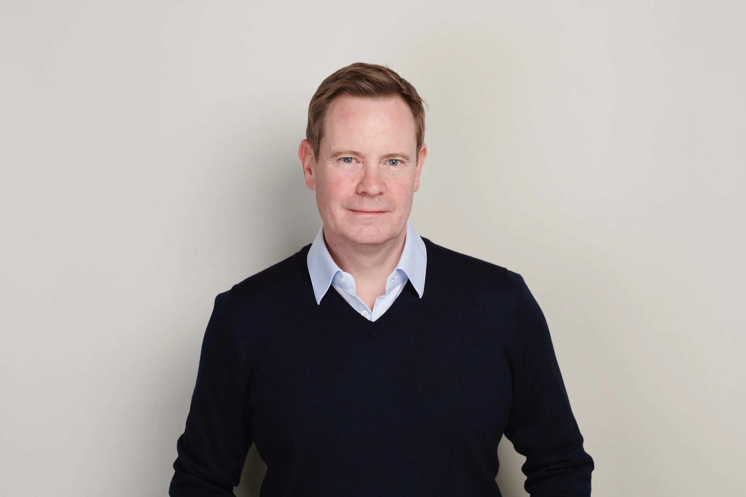 A man with red hair and blue eyes wearing a light blue collared shirt under a black sweater, standing against a plain light-colored background.
