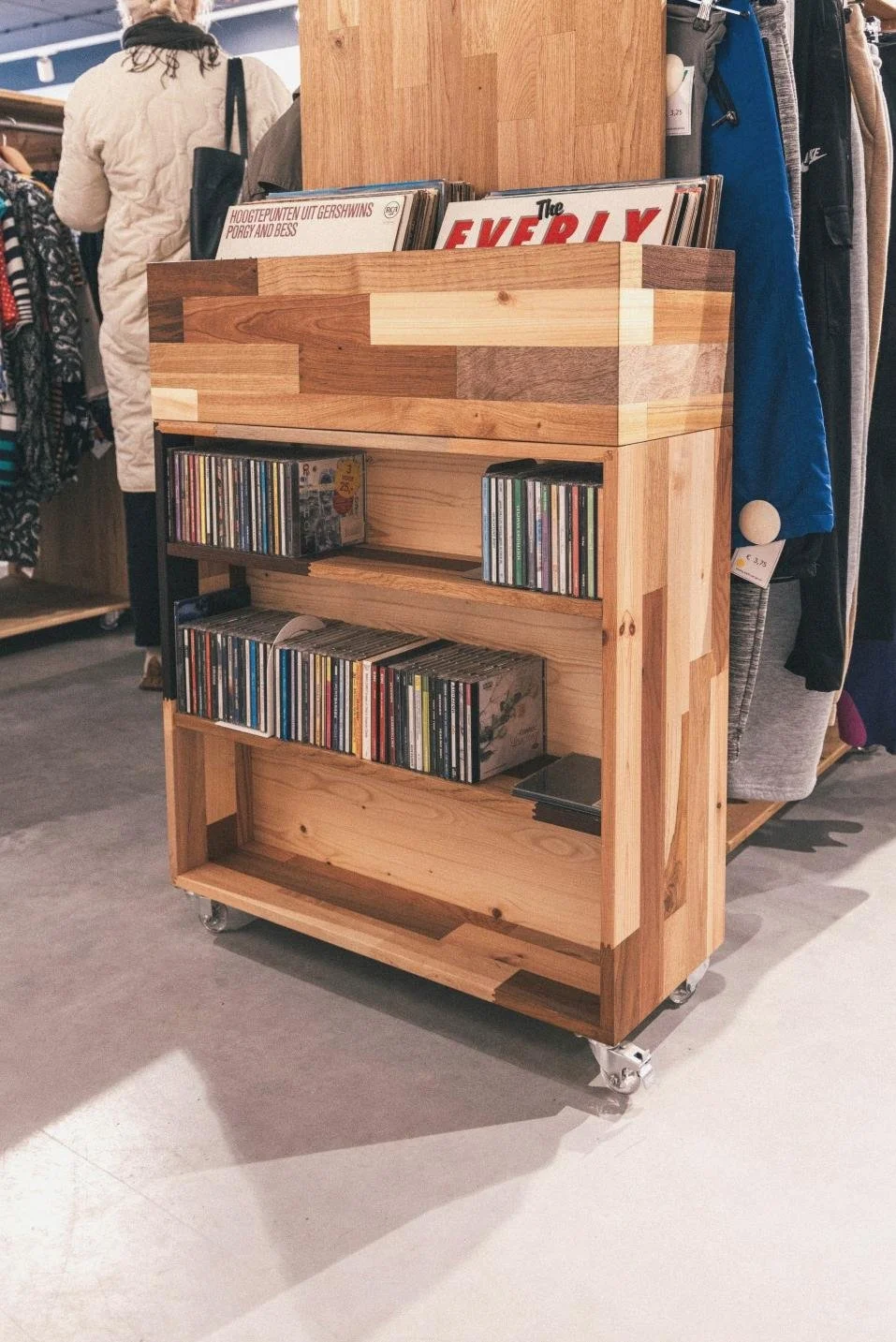 Een houten platenkast met wieltjes gevuld met vinylplaten, omringd door kledingrekken in een warenhuis of tweedehandswinkel.