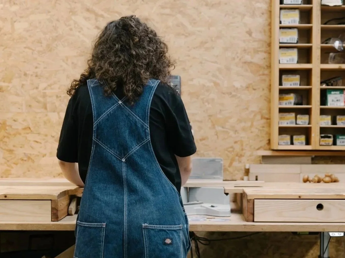 A person with curly hair wearing a denim apron standing at a woodworking workbench in a workshop.