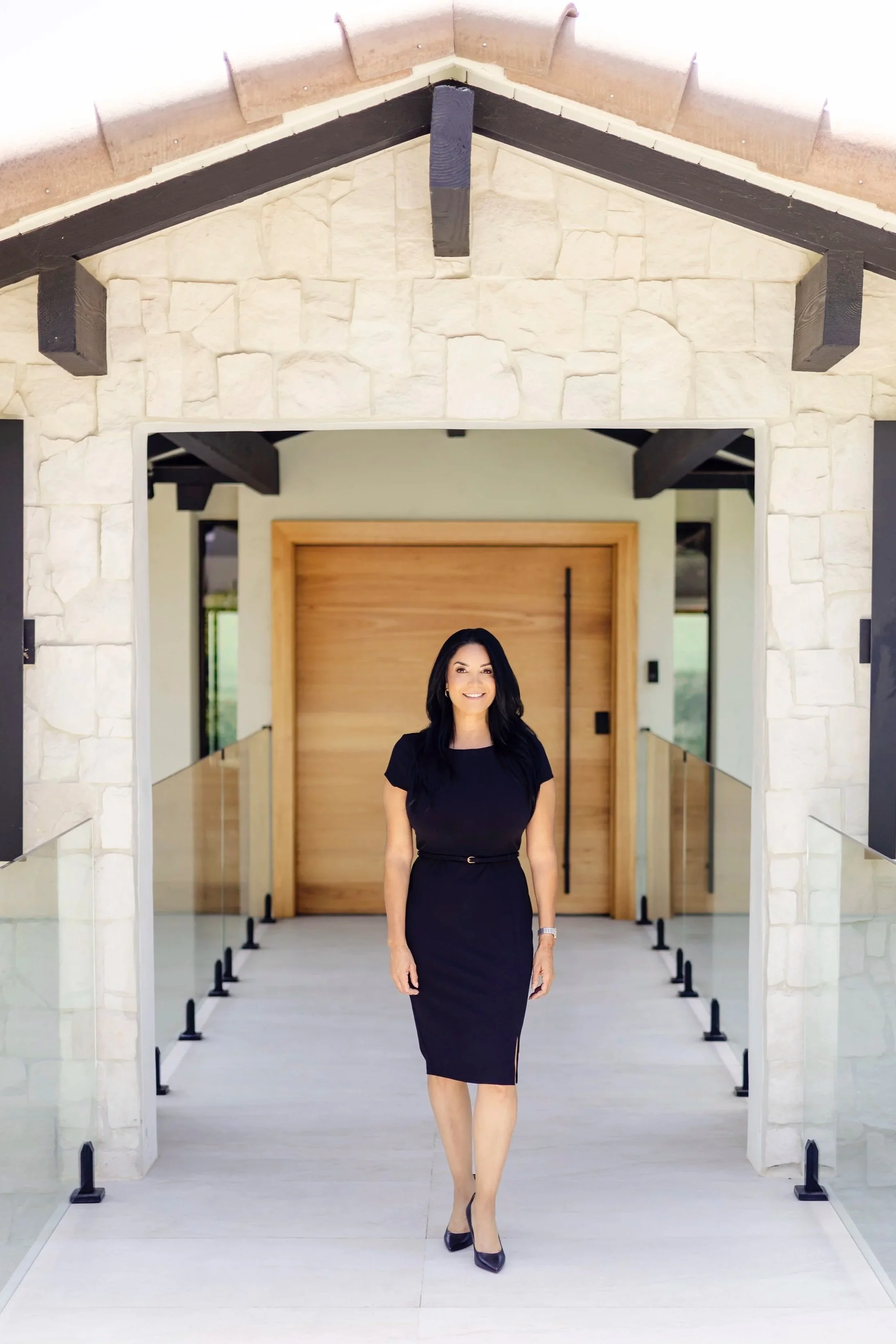 A woman in a black dress walking through a modern building with stone and wood accents, smiling.