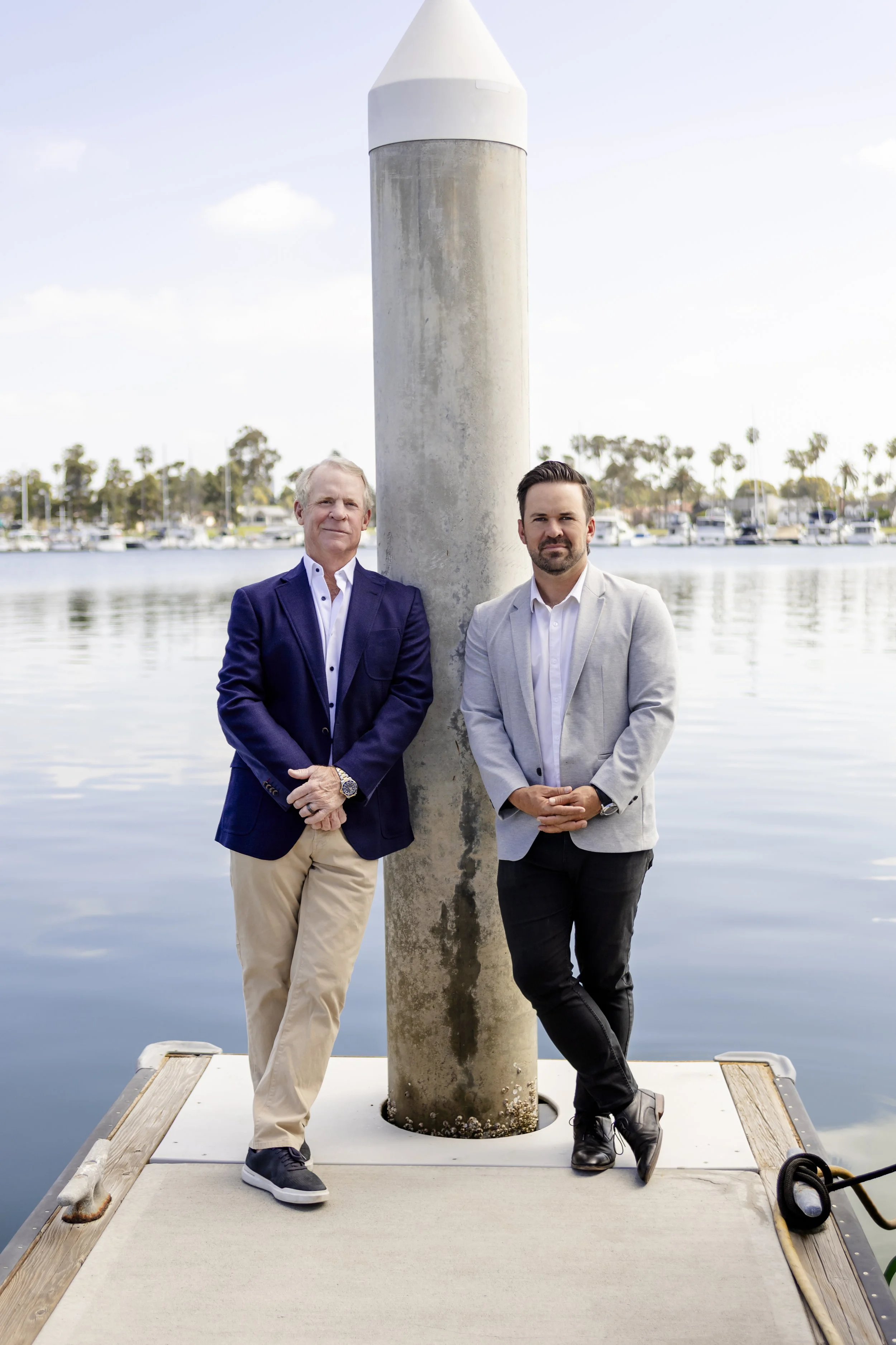 Two men in suits standing on a dock by the water, next to a large, cylindrical concrete structure, with a marina and palm trees in the background.