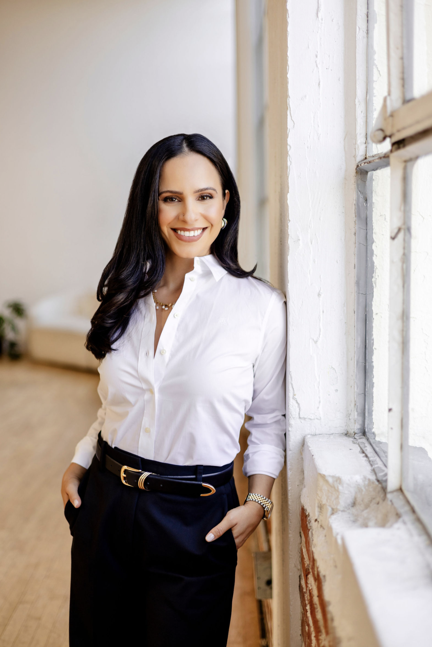 A woman with long dark hair, wearing a white button-up shirt and dark pants with a black belt, standing by a window with her hand in her pocket, smiling at the camera indoors.