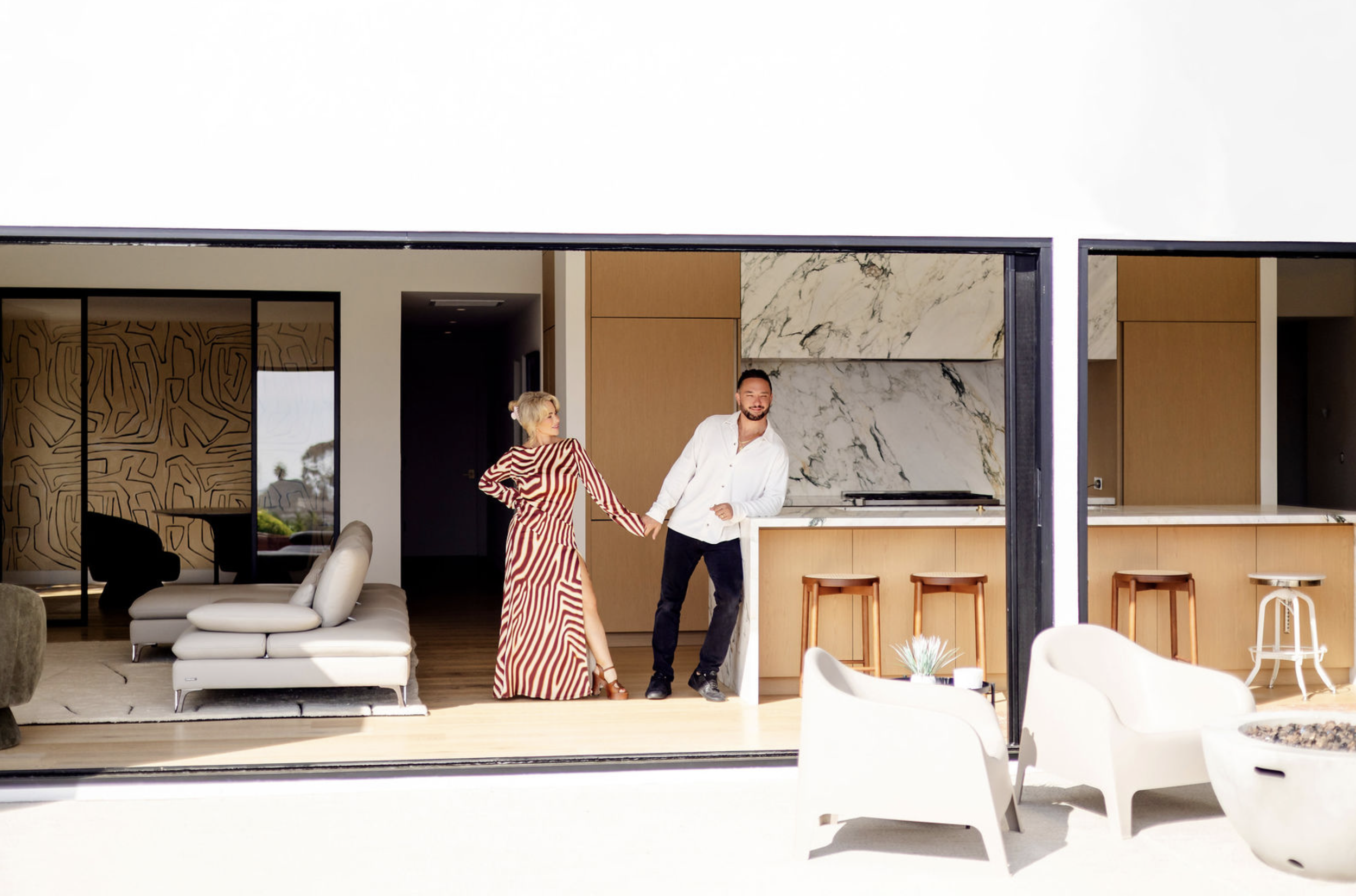 A man and woman holding hands inside a modern home, with the woman wearing a red and white striped dress and the man in a white shirt and dark pants. The interior features a white sofa, marble walls, and a kitchen area with wooden cabinets and bar stools.