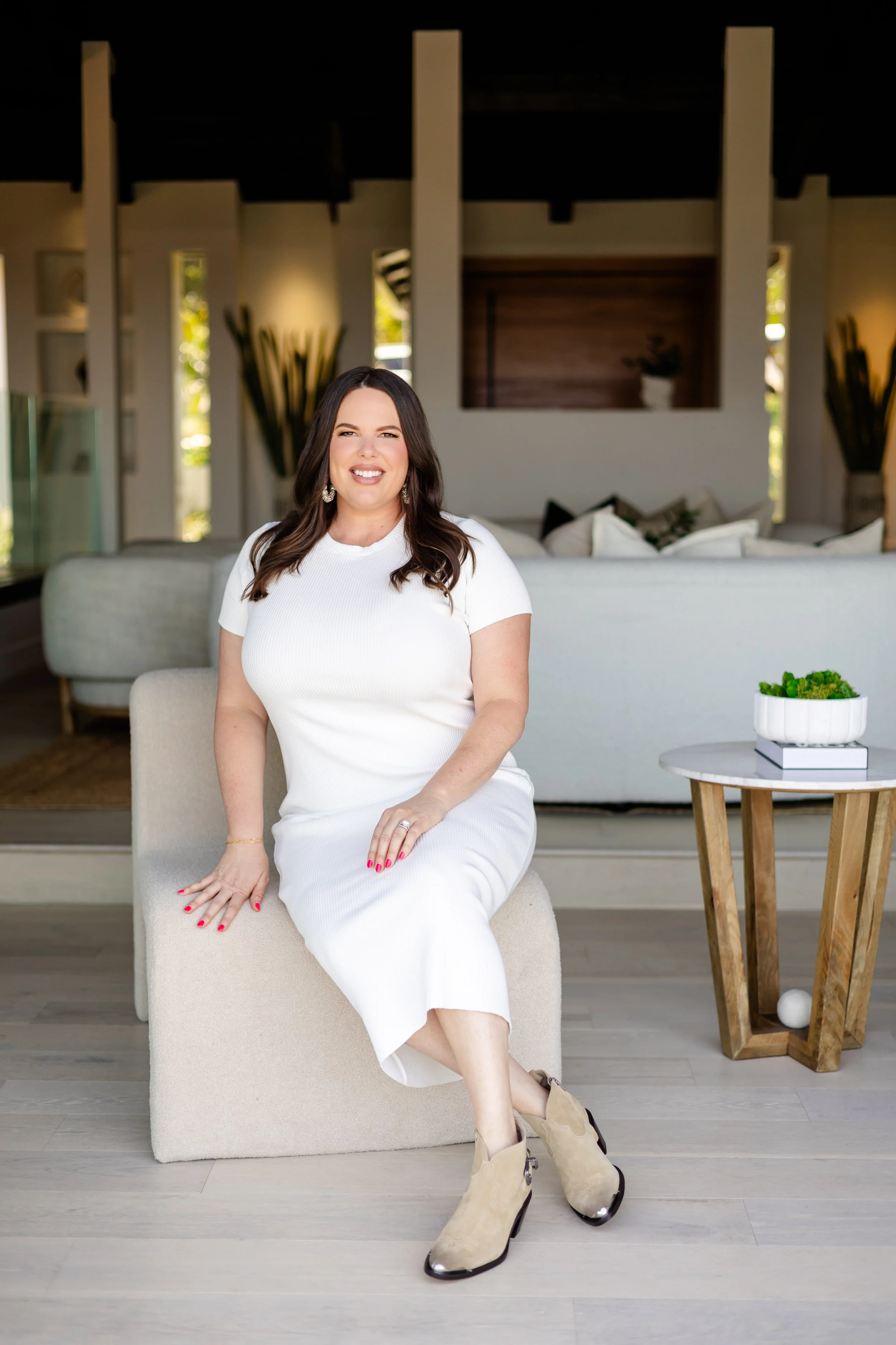 Smiling woman sitting on a beige chair in a modern living room, wearing a white dress and beige boots, with a small round table holding a plant nearby.