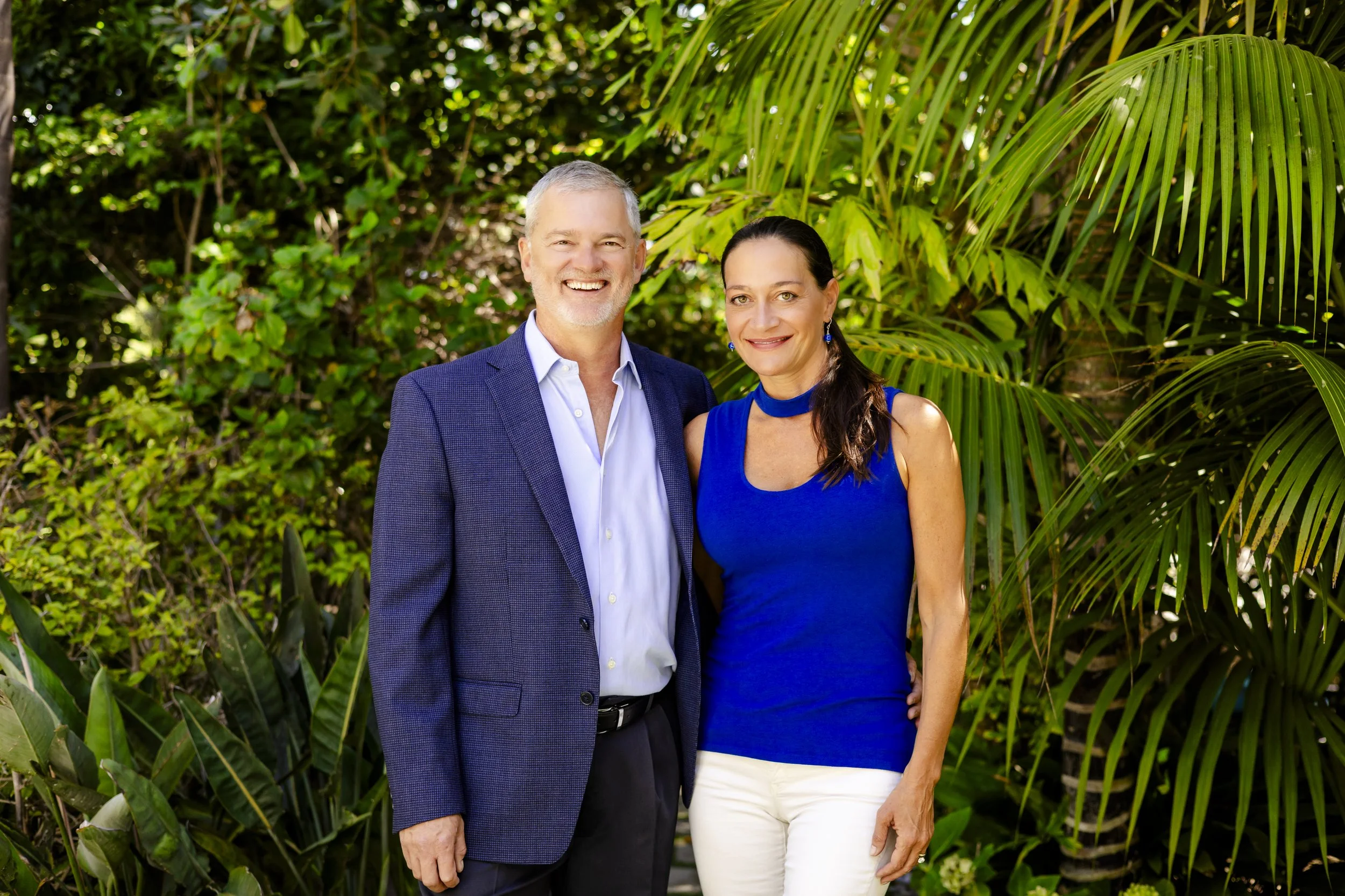 A smiling middle-aged man and woman standing together outdoors in a lush green tropical garden.