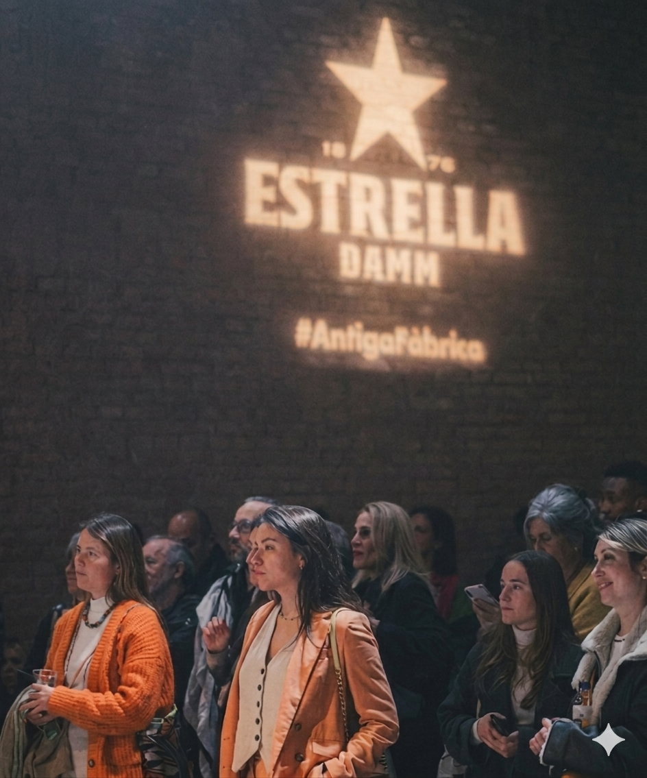 People attending an event in a dimly lit room with a large projected logo on a brick wall that reads 'Estrella Damm' with a star above it, and the hashtag '#AntigaFabrica' below.