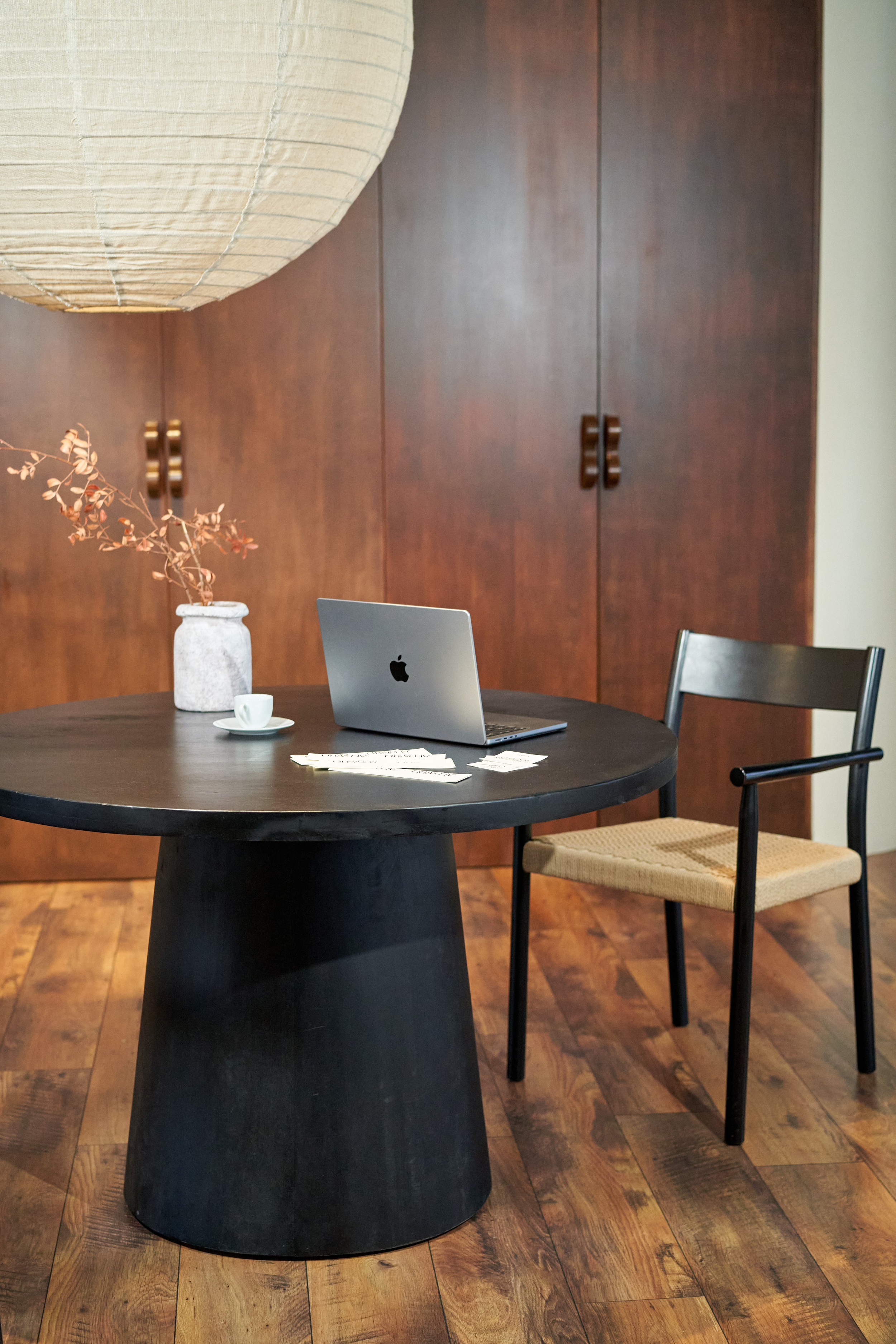 A black oval table with a silver MacBook, a white vase with dried branches, a small cup, and some papers. A black chair with a woven seat is beside the table. Behind, there are wooden cabinets.