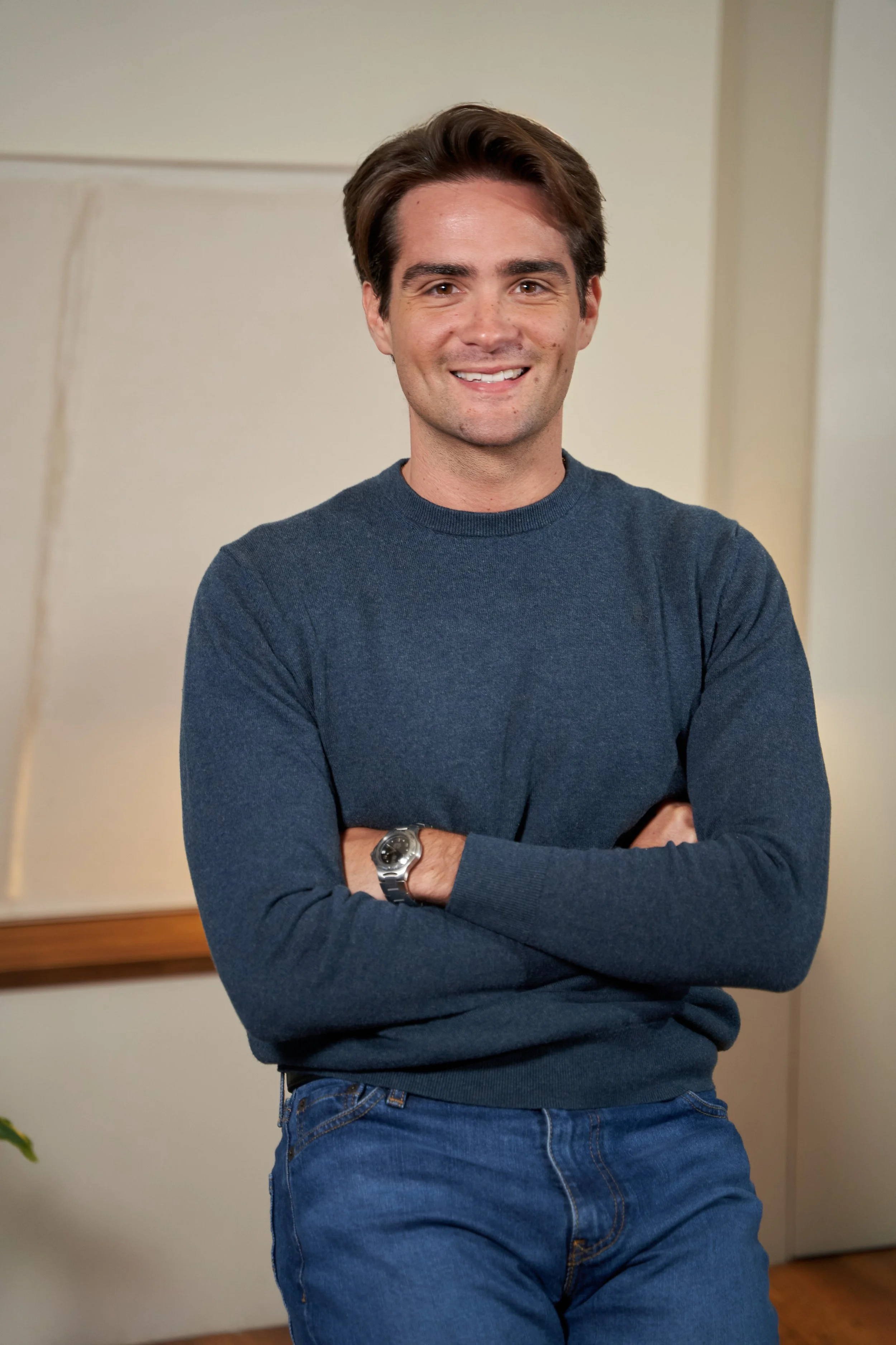 A smiling young man with brown hair, wearing a dark gray sweater and blue jeans with arms crossed, standing in a room with neutral beige walls.