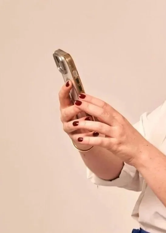 A woman with dark red nail polish is holding a smartphone with a clear case against a plain, light-colored background.