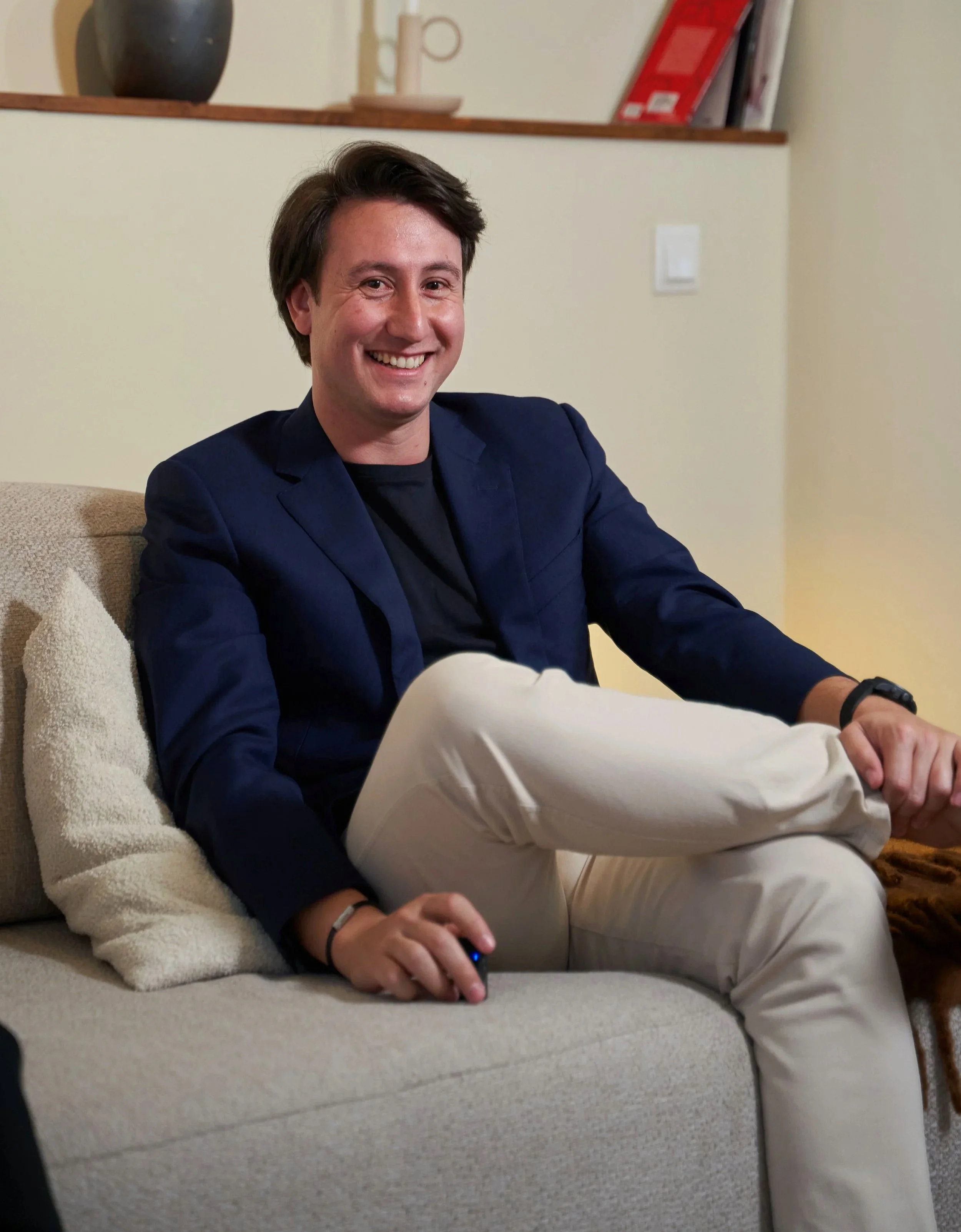 A man sitting on a beige couch, smiling, wearing a dark blazer, black shirt, and beige pants, holding a remote control in his right hand.