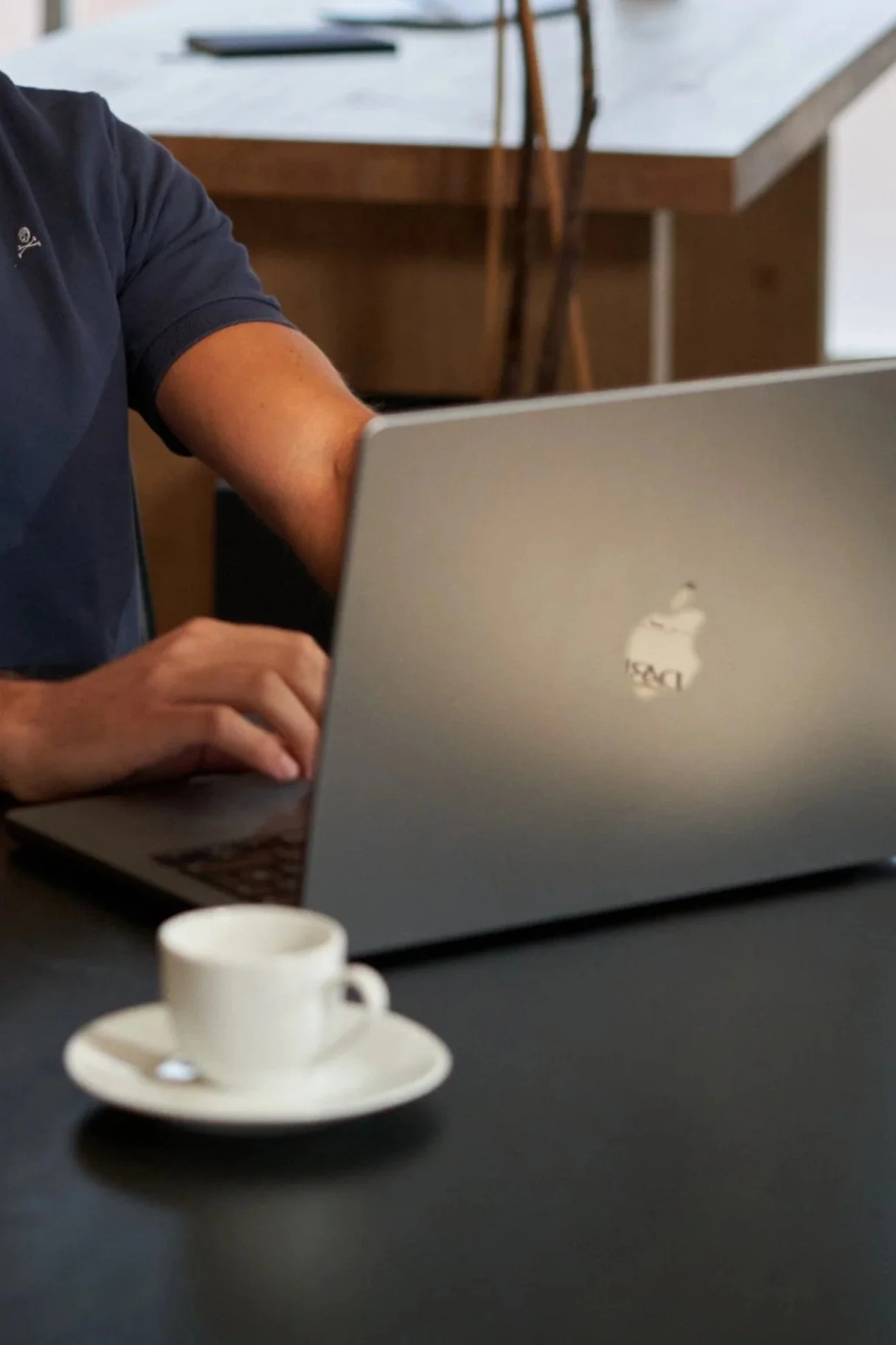 Person working on a silver MacBook laptop at a black table, with a white coffee cup and saucer in front, inside a modern room with wooden furniture.