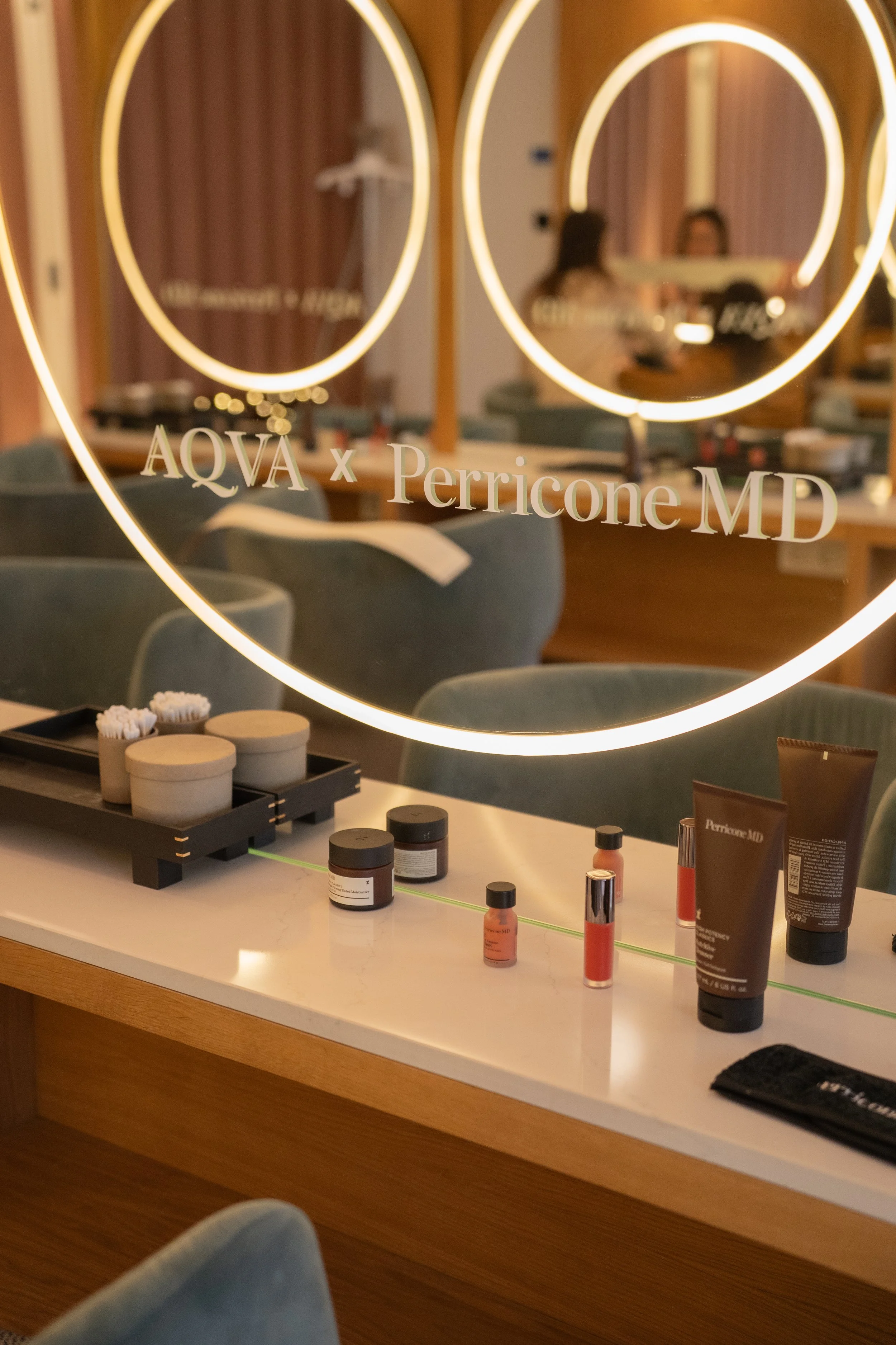A mirror with neon lights around its border, reflecting a room with a table having skincare or beauty products, and two people sitting at a counter in the background.