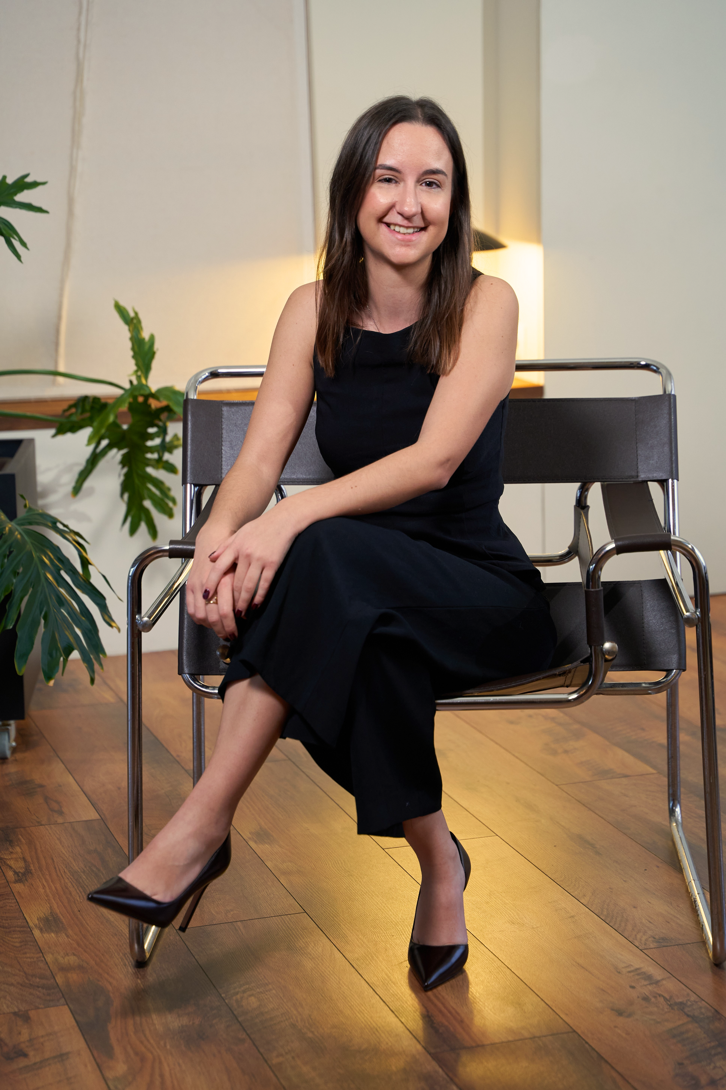 A woman wearing a black sleeveless dress and black high heels sitting on a modern chair with metallic arms in a room with wooden flooring, a green plant, and warm lighting.