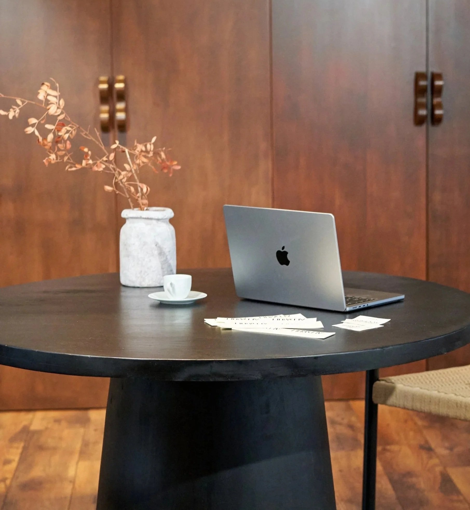A round black table with a MacBook, a small white cup and saucer, a gray vase with dried branches, and some papers, set against a wooden wall with cabinet doors.