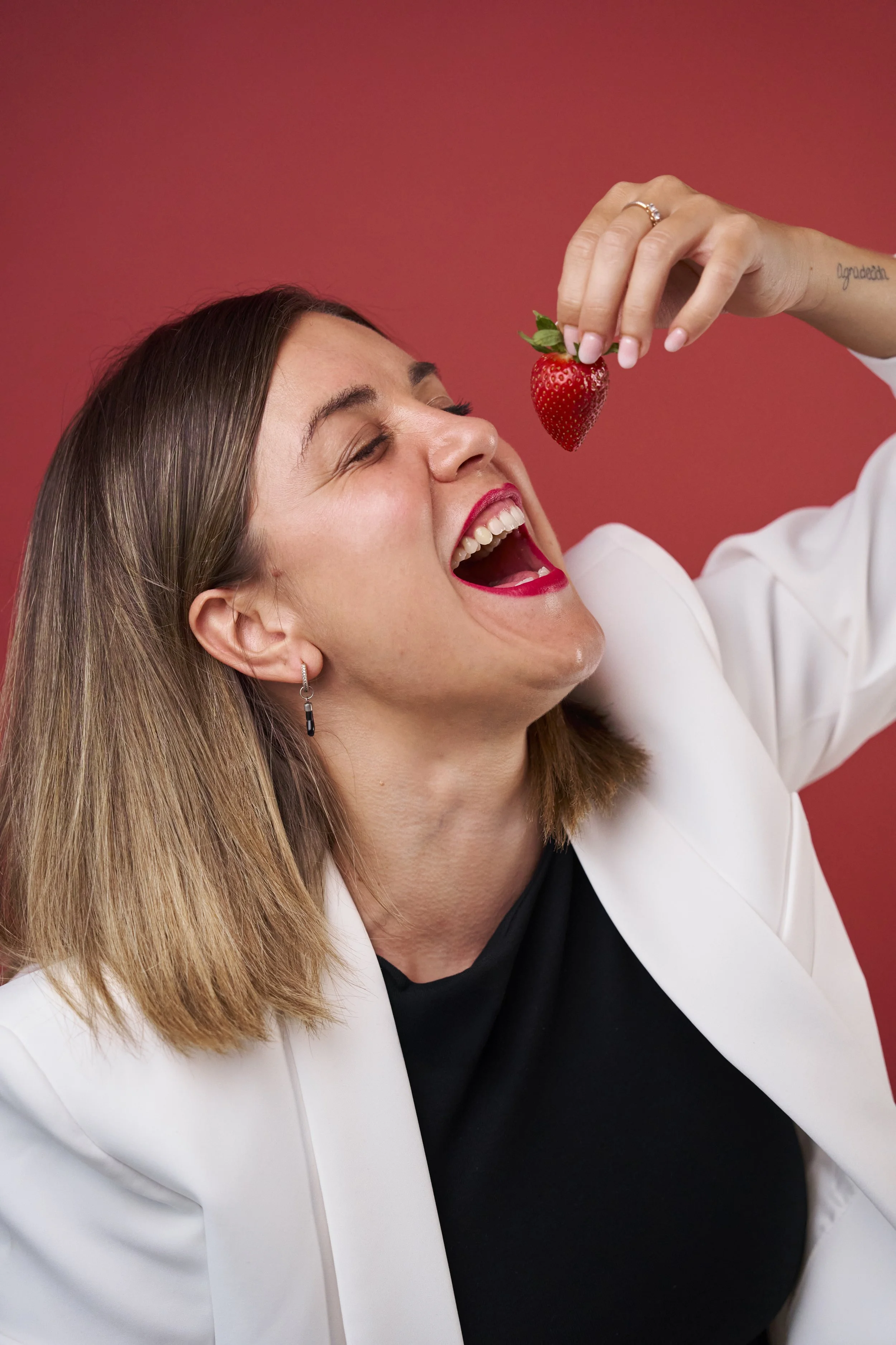 A woman with shoulder-length hair, wearing a white blazer and black top, is smiling with her mouth open as she is about to eat a strawberry held above her mouth against a red background.