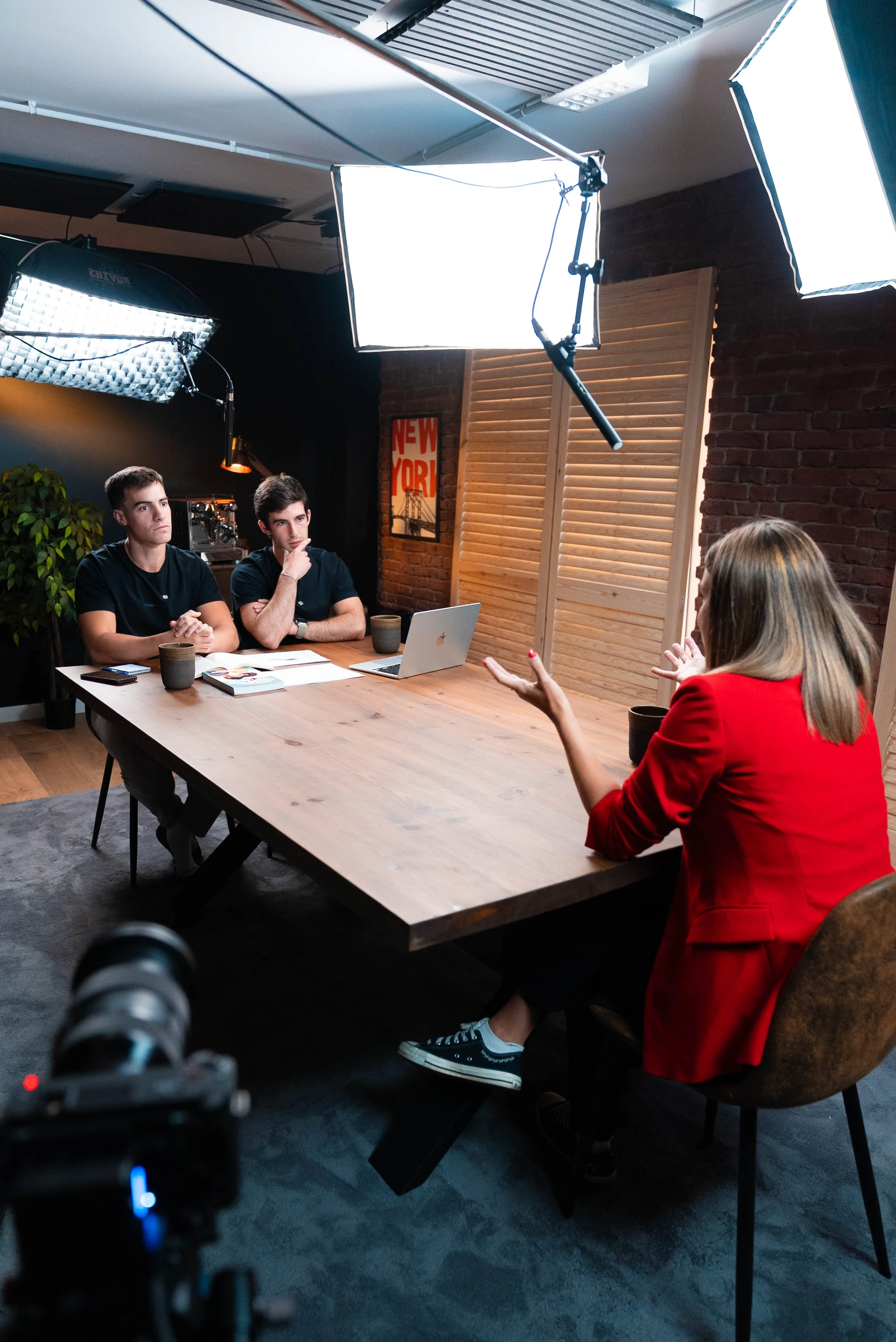 A woman in a red blazer is conducting an interview or discussion with two young men seated at a wooden table in a studio setting. The scene includes professional lighting equipment overhead, a camera in the foreground, and a brick wall with a 'New York' poster in the background.