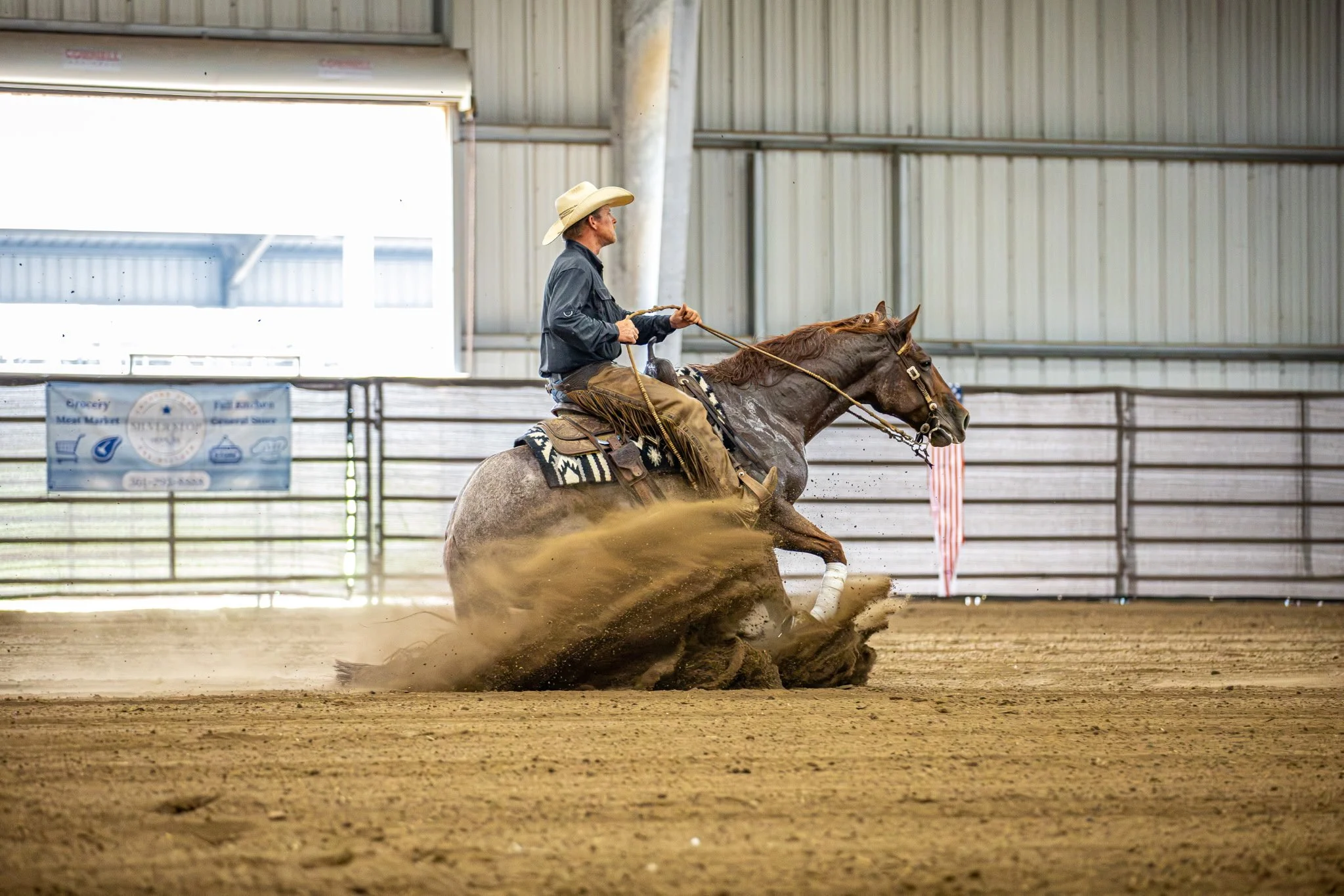 A cowboy wearing a wide-brimmed hat, sunglasses, and jeans rides a horse in an indoor arena with a crowd watching in the background.