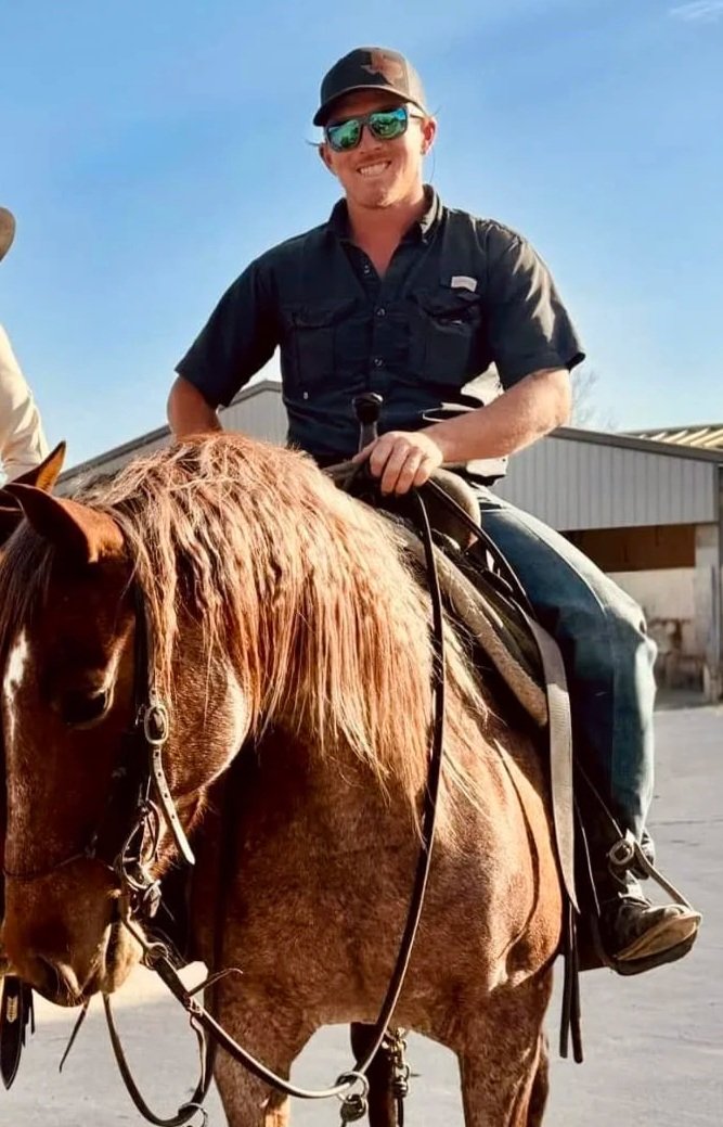 A cowboy wearing a wide-brimmed hat, sunglasses, and jeans rides a horse in an indoor arena with a crowd watching in the background.