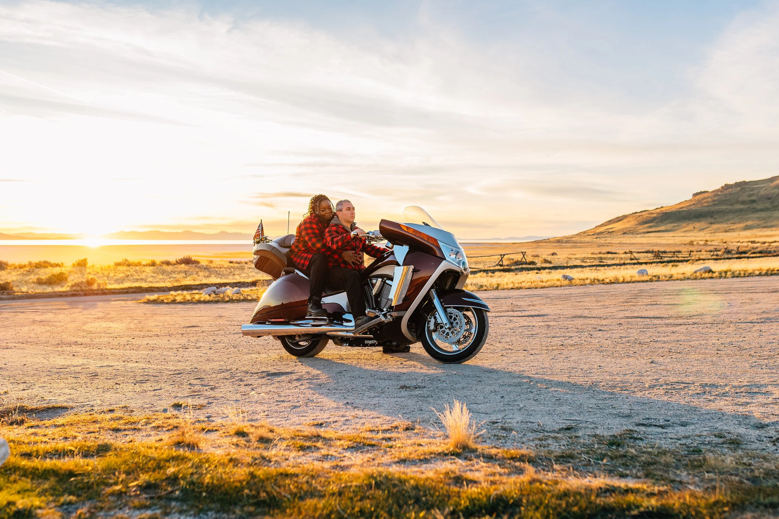 Two people on a motorcycle riding through a desert landscape at sunset.