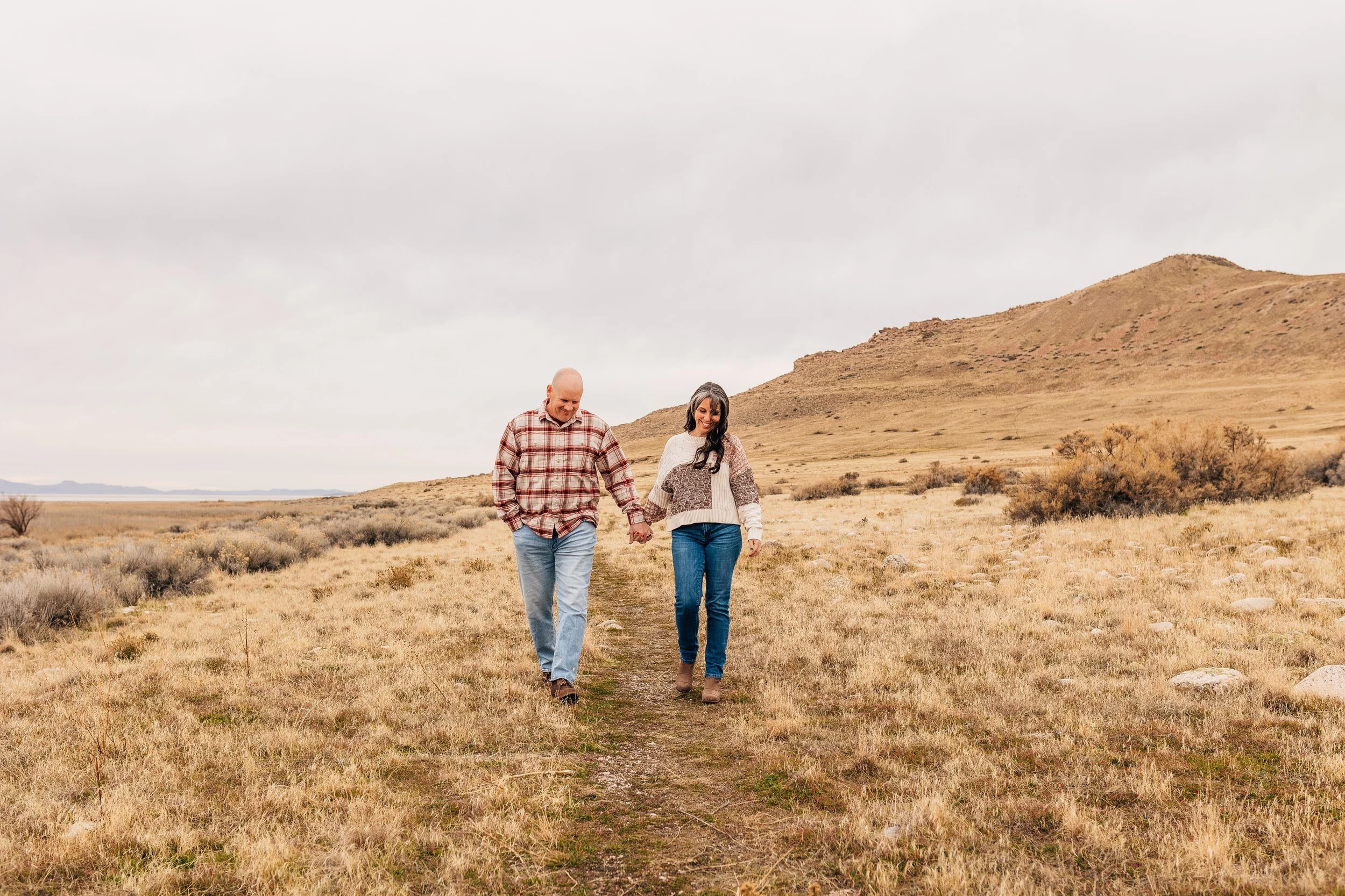 A couple walking hand in hand on a dirt path in a dry, grassy landscape with hills in the background during overcast weather.