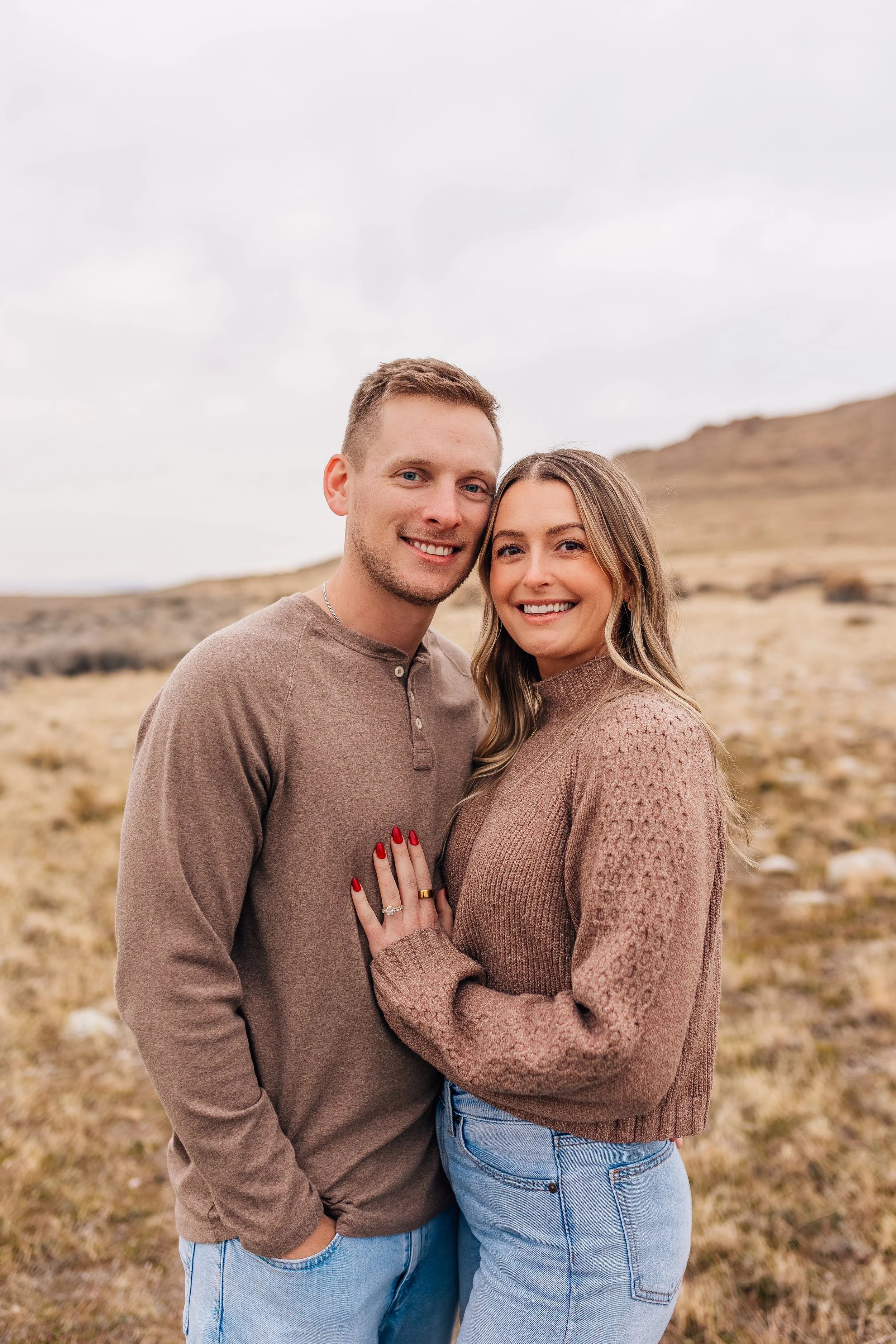 A young couple stands outdoors in a grassy, hilly area with overcast skies. The man has short, light brown hair and is wearing a brown long-sleeve shirt. The woman has long, light brown hair and is wearing a knitted brown sweater, light blue jeans, a