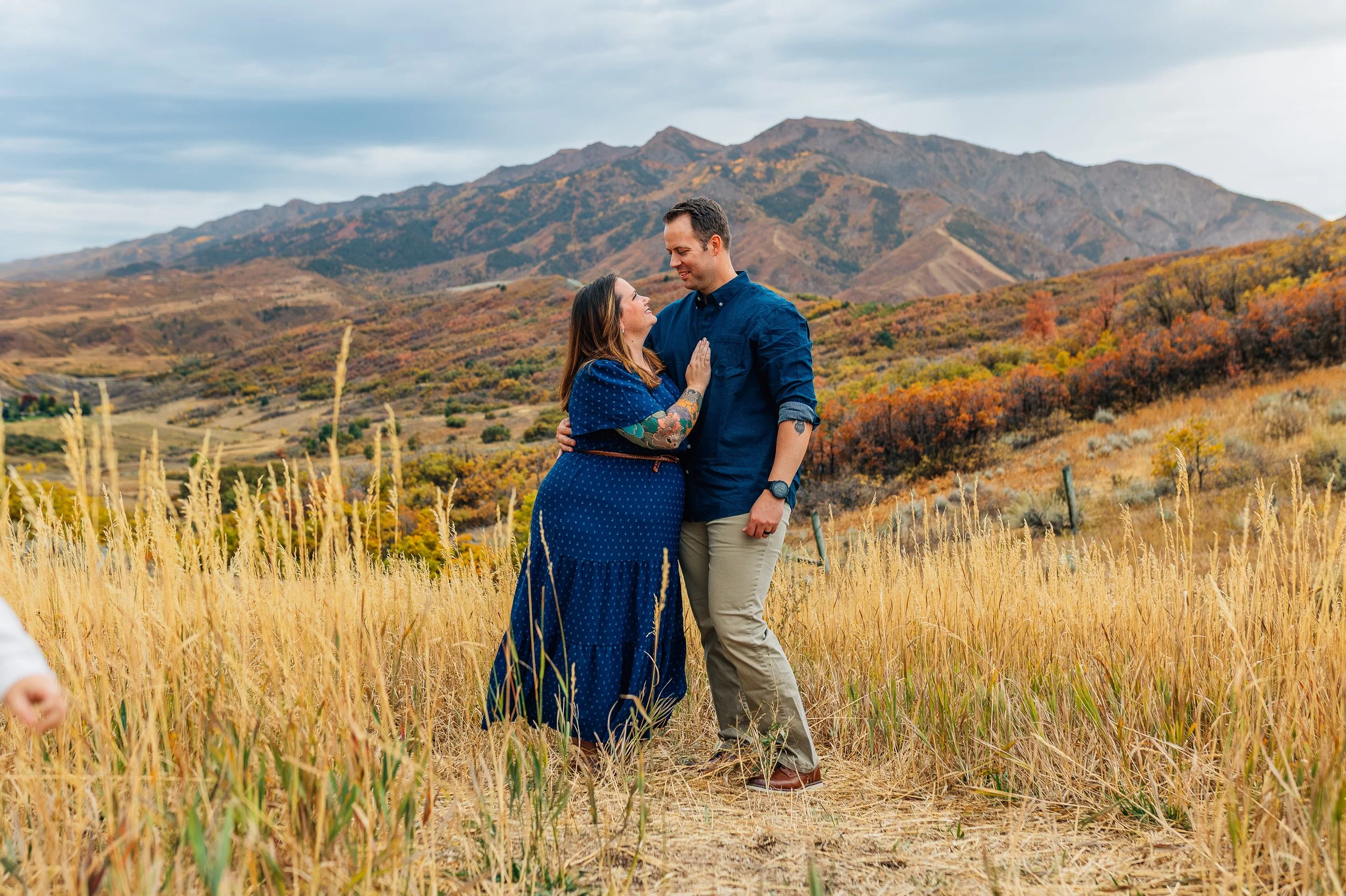 A couple stands close together in a field of tall, yellow grass, with mountains and autumn foliage in the background. The woman is wearing a blue dress and has tattoos on her arms, while the man is wearing a navy blue shirt and beige pants. They are 