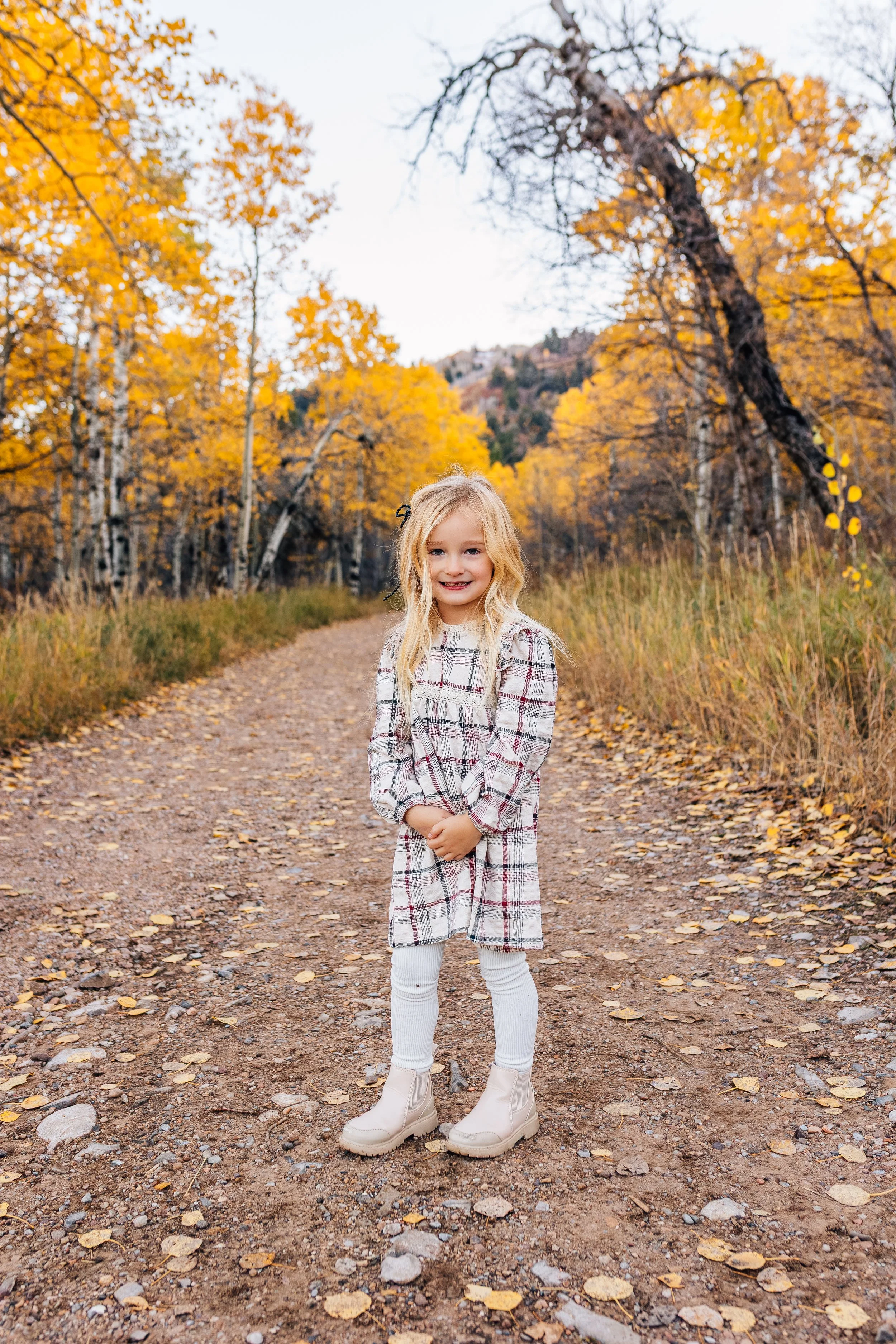 A young girl with blonde hair in a checkered dress and white boots standing on a dirt path in a forest with autumn yellow leaves.