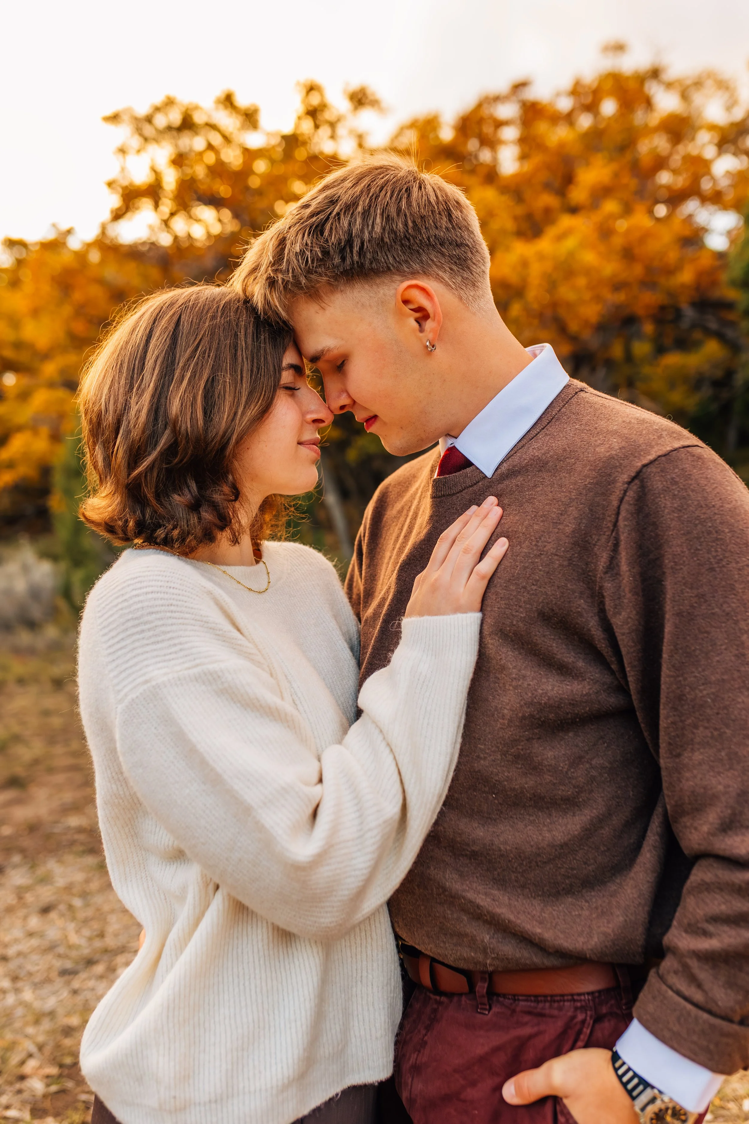 A young couple with foreheads touching, eyes closed, in an outdoor setting during autumn with orange and yellow foliage in the background.