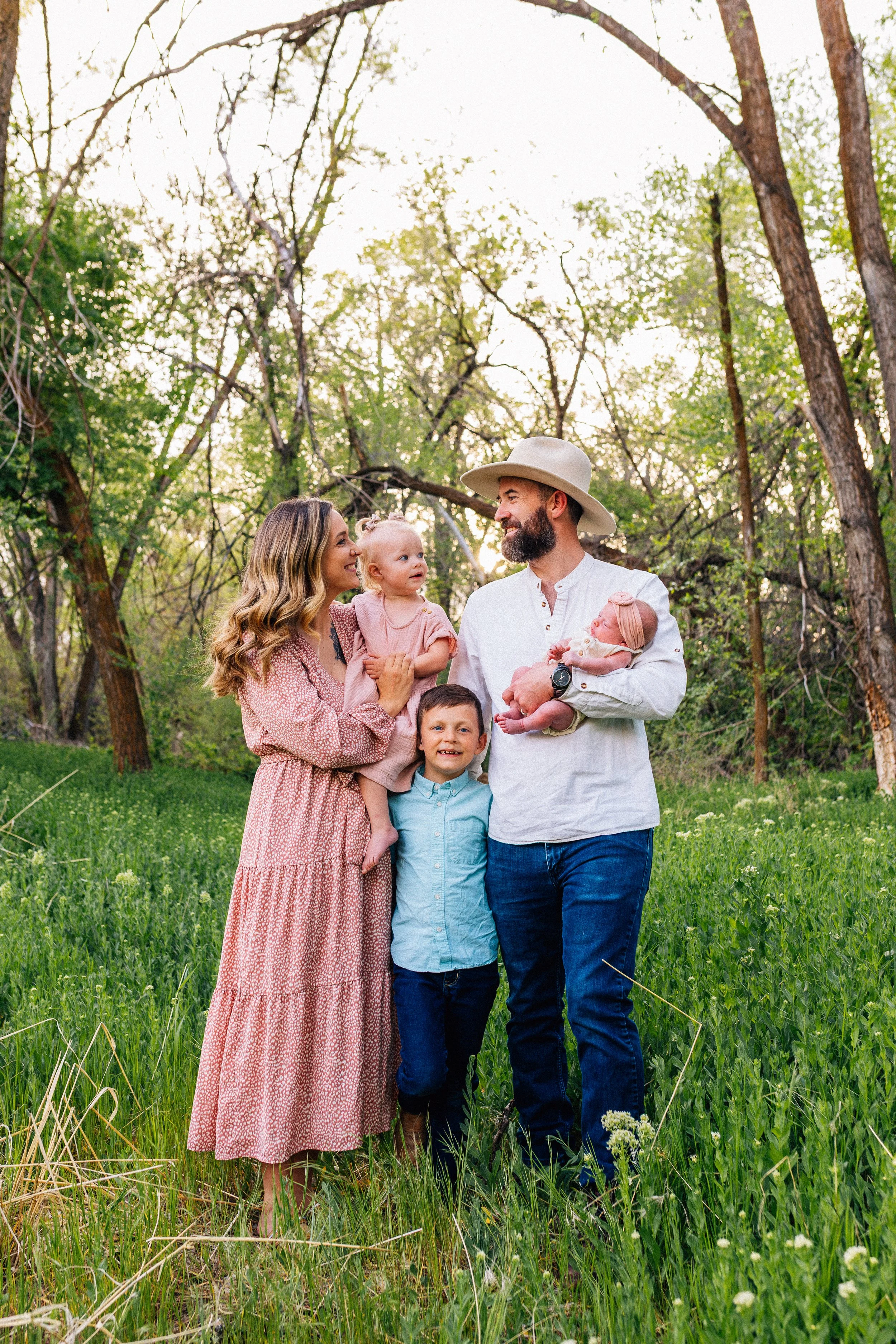A family of five outdoors in a wooded area during daytime. The woman holds a young girl, and the man holds a newborn baby. A young boy stands in front of them, all smiling and facing each other.