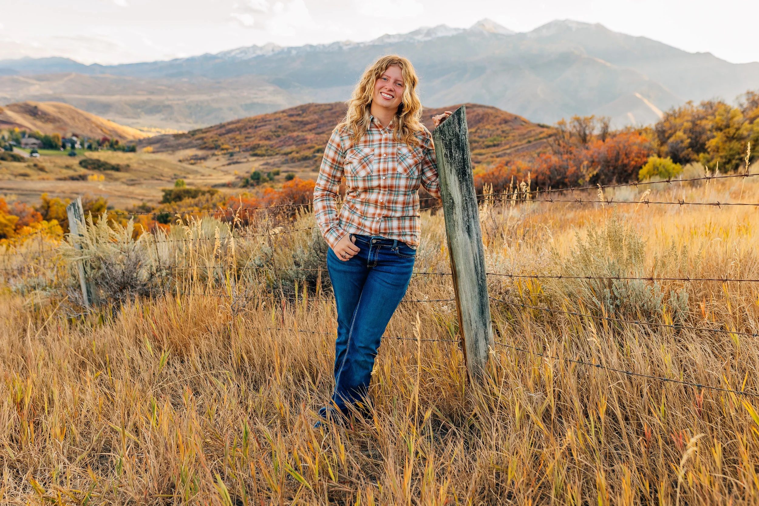 A woman standing next to a wooden fence in a grassy field with mountains and colorful autumn trees in the background.