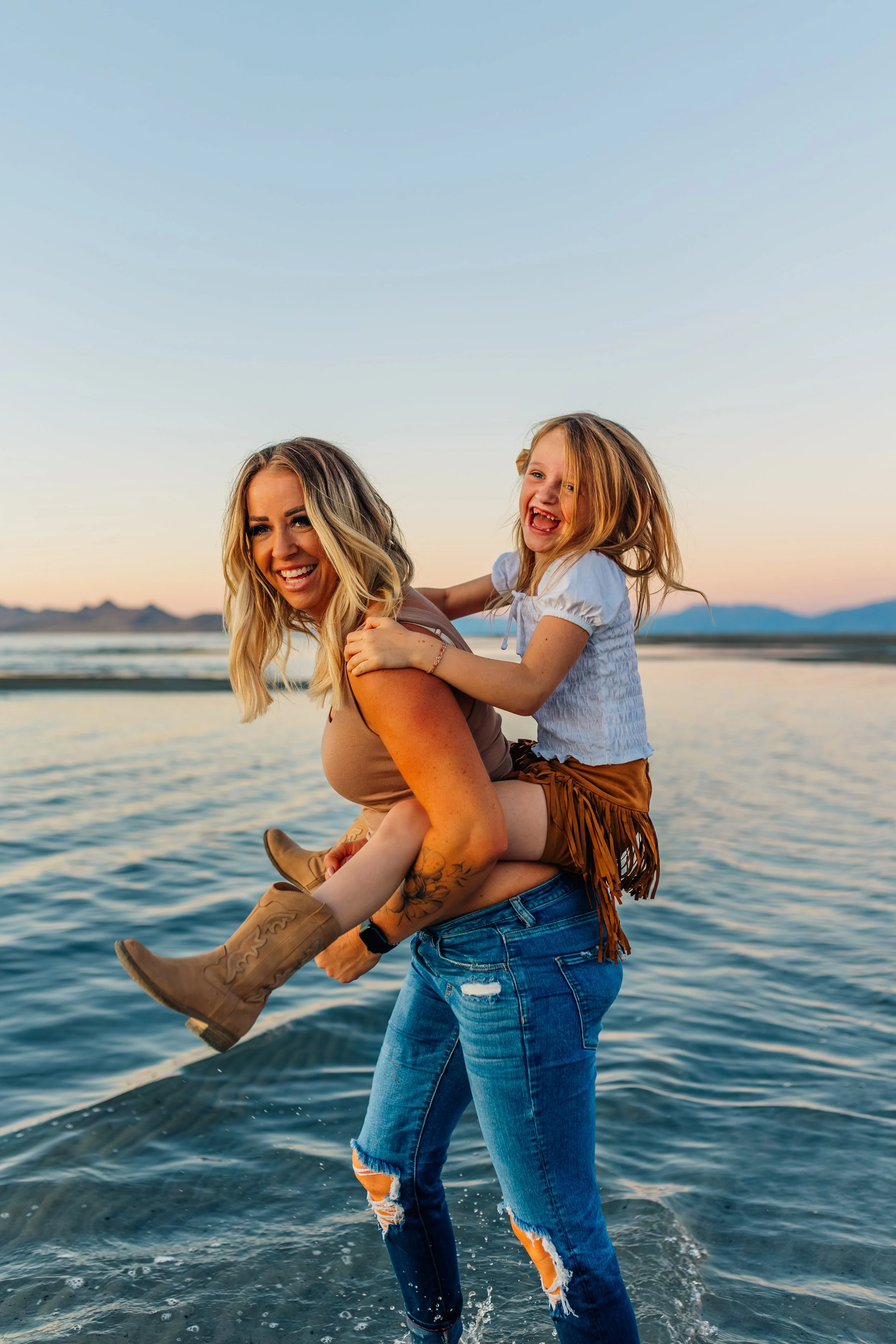 A woman carrying a girl on her shoulders by the water at sunset.