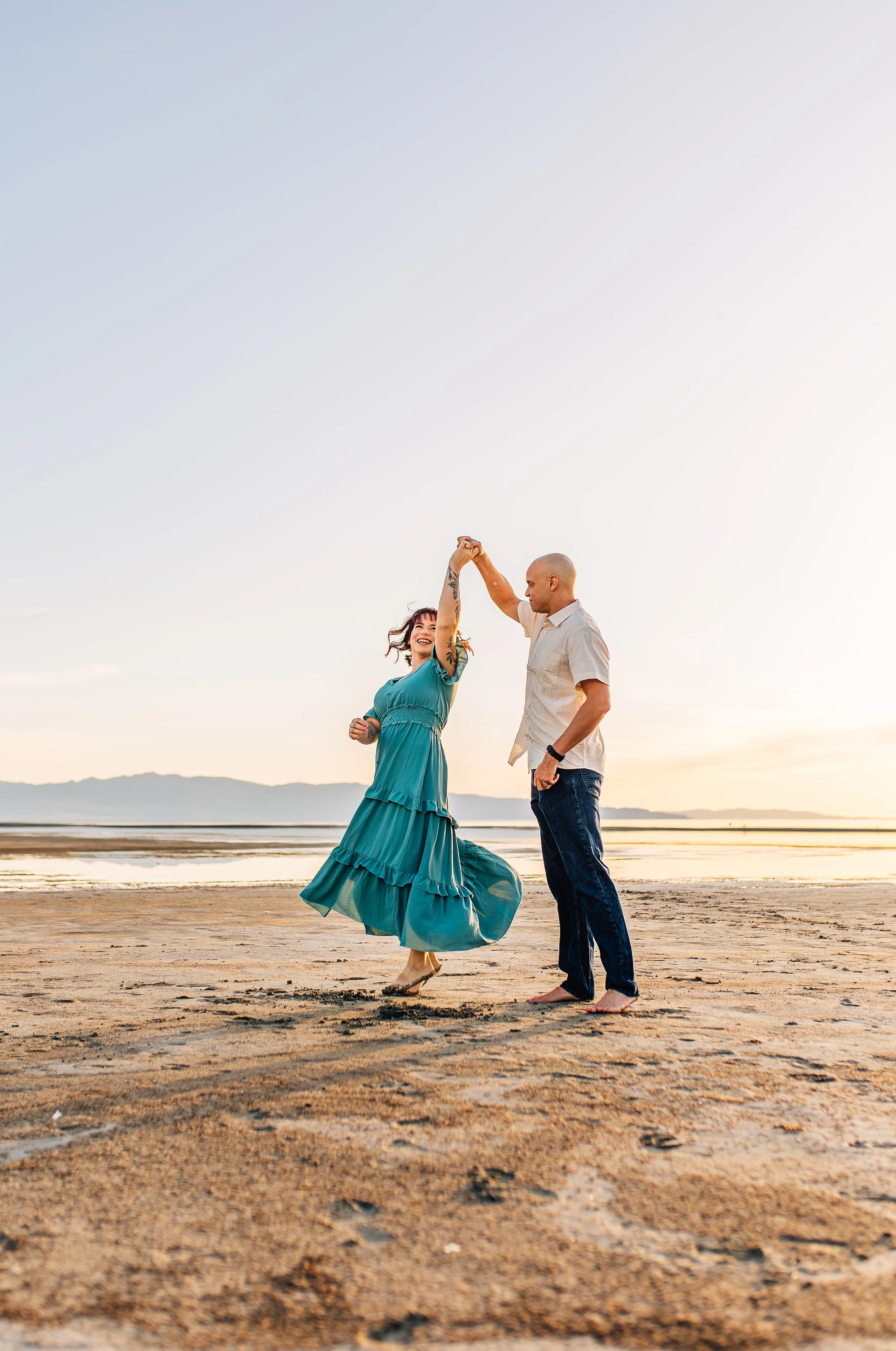 A couple dancing on the beach during sunset, with the woman wearing a flowing teal dress and the man in a white shirt and jeans, holding hands and smiling.