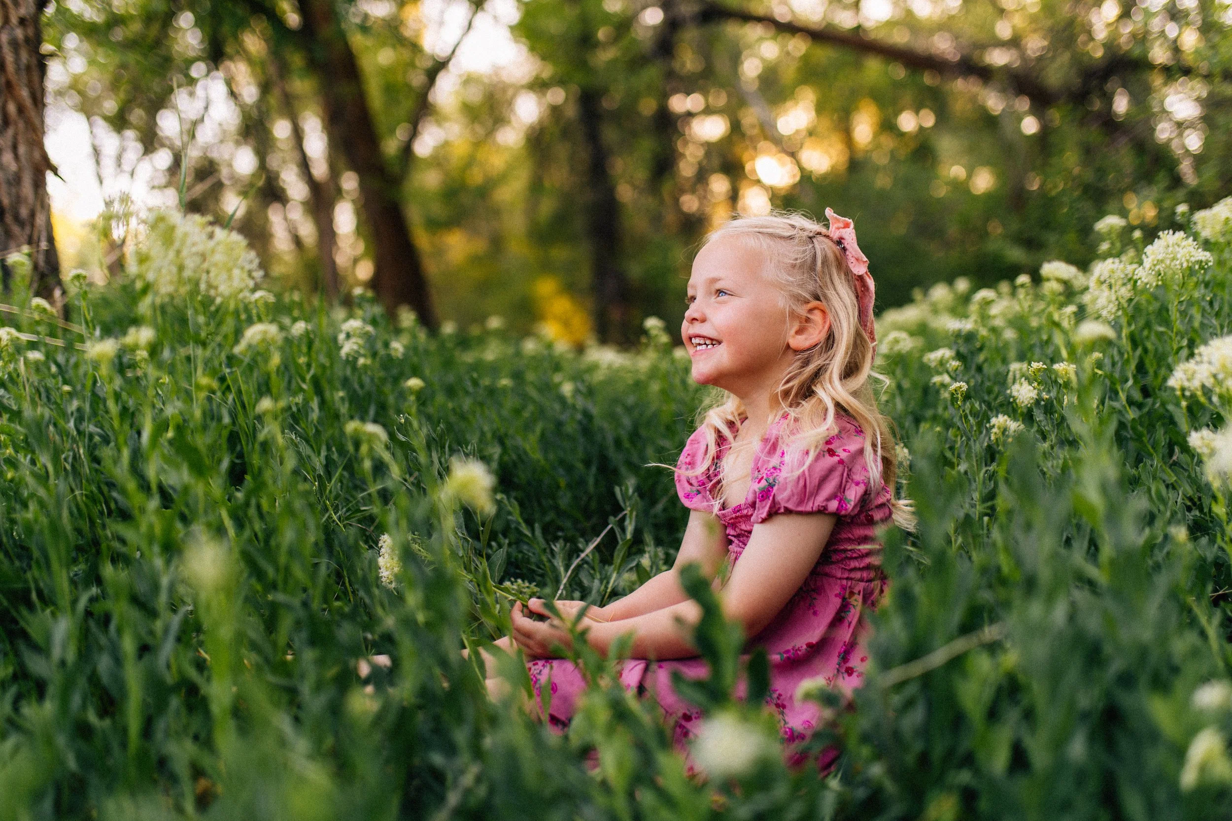 A young girl with blonde hair in a pink dress sitting among white flowers in a green garden or forest, smiling and looking to the right.