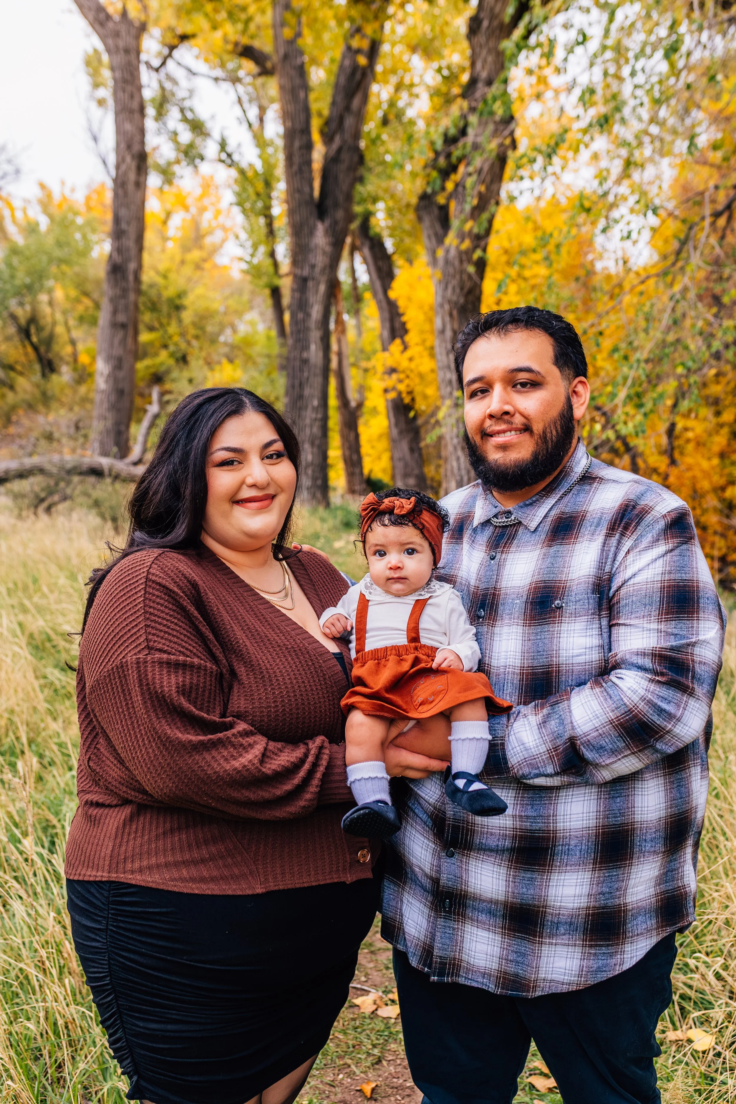 A family of three outdoors in a wooded area during fall, consisting of a woman with long dark hair, a man with a beard, and a baby girl dressed in autumn clothing, all smiling at the camera.