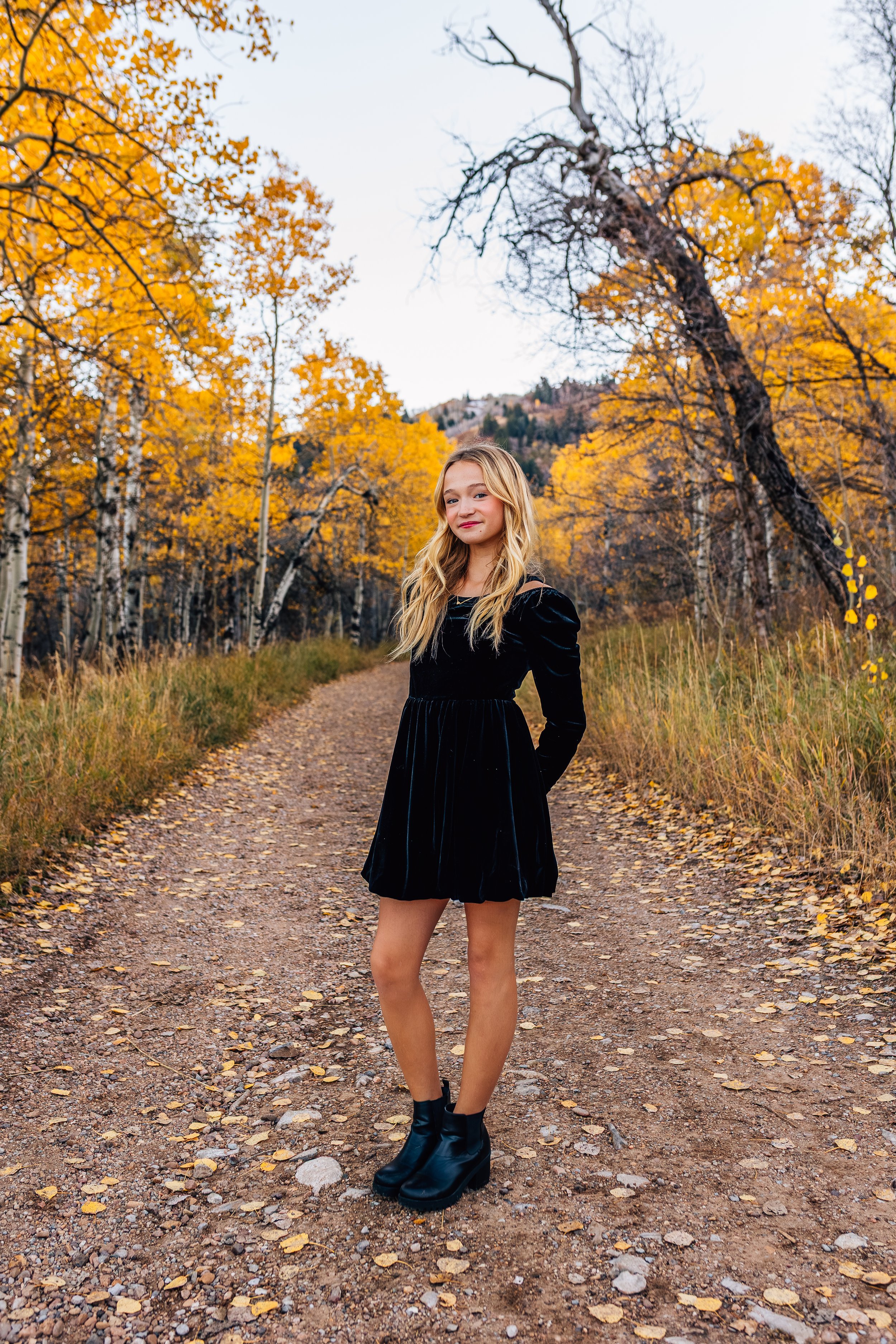 A young woman with long blonde hair standing on a dirt path in an autumn forest with yellow and orange leaves.