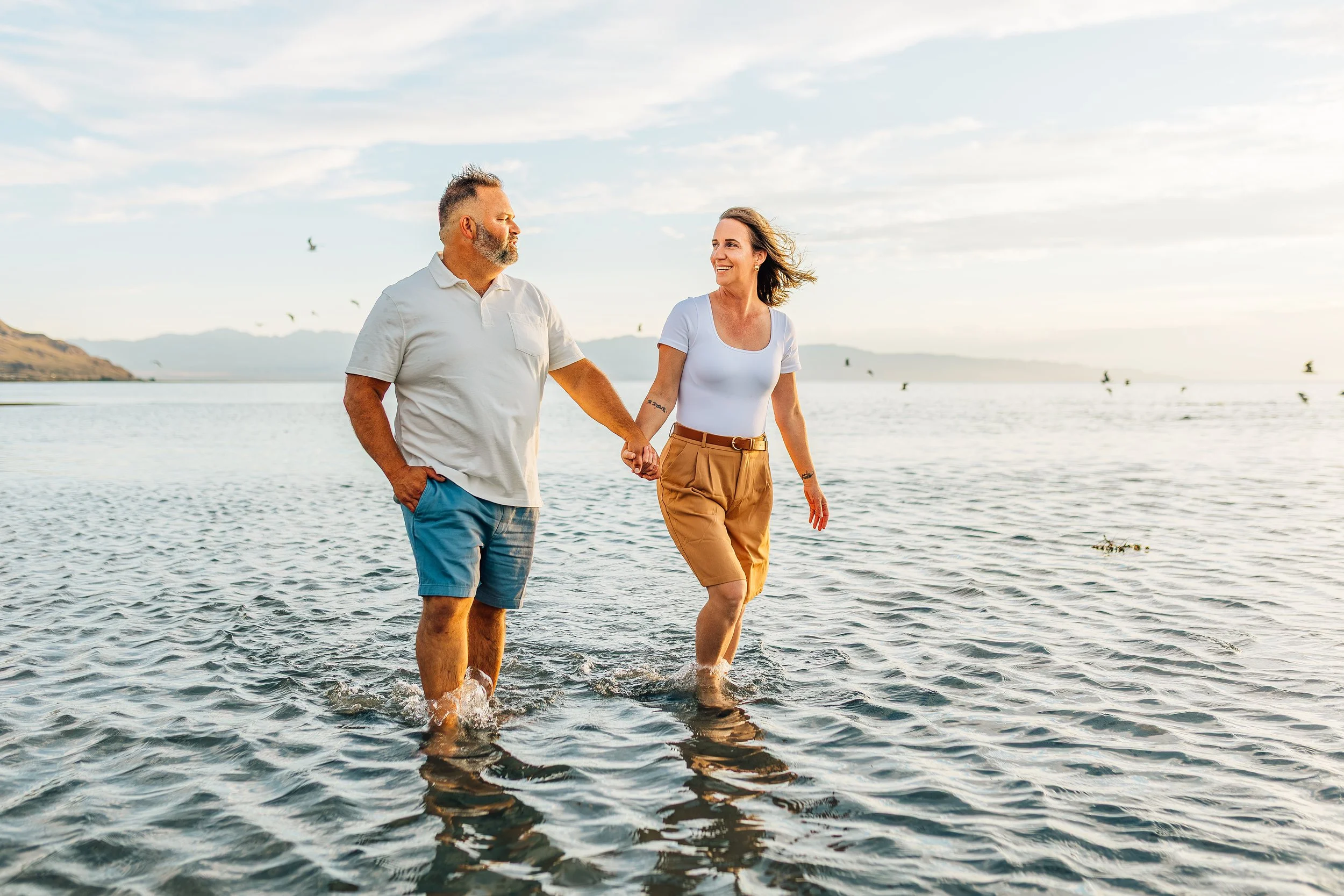 A couple holding hands and walking in shallow water at the beach during sunset, smiling and enjoying the moment.