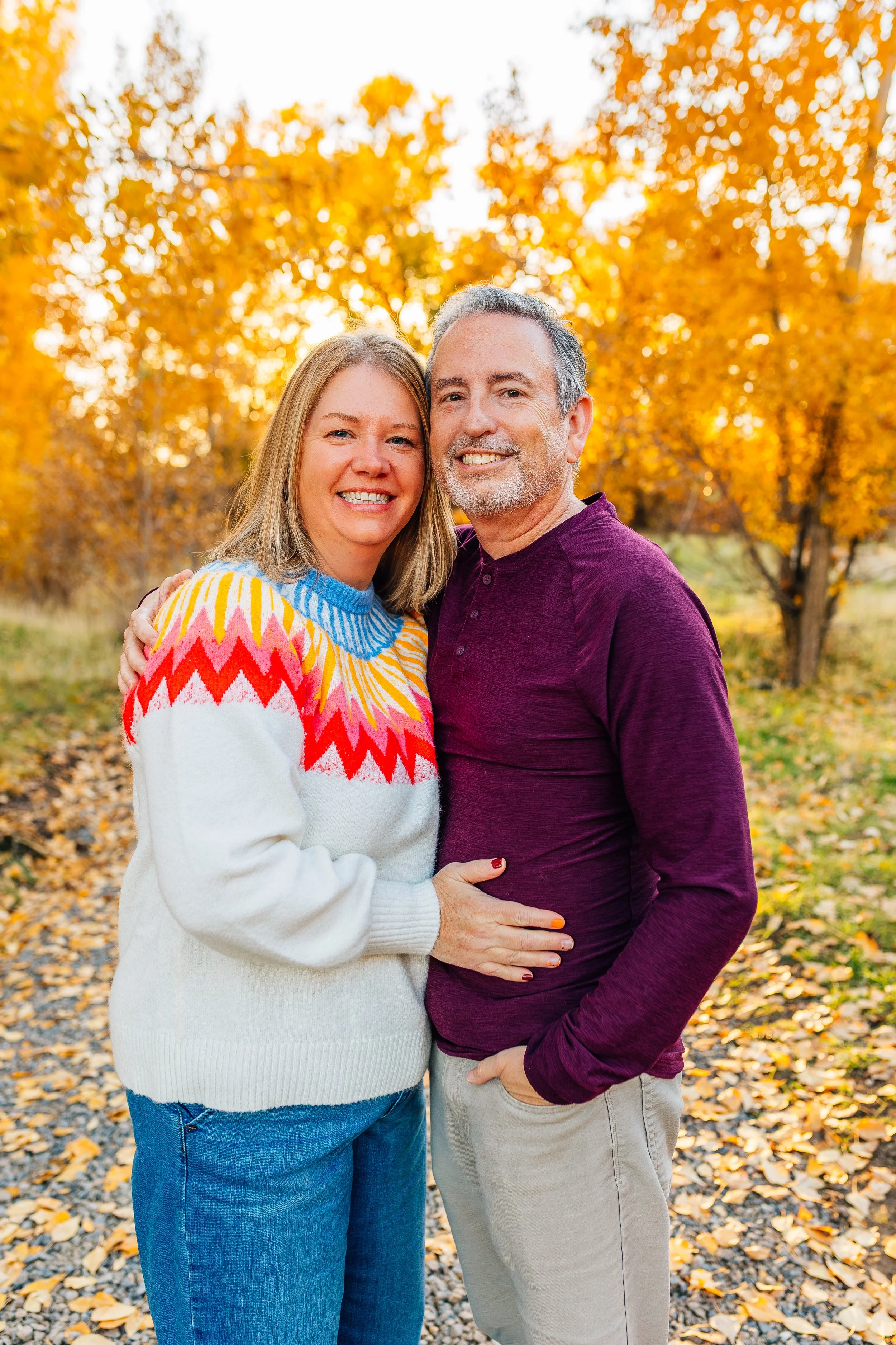A smiling middle-aged couple standing close together outdoors during autumn, surrounded by trees with orange leaves, wearing warm clothes.