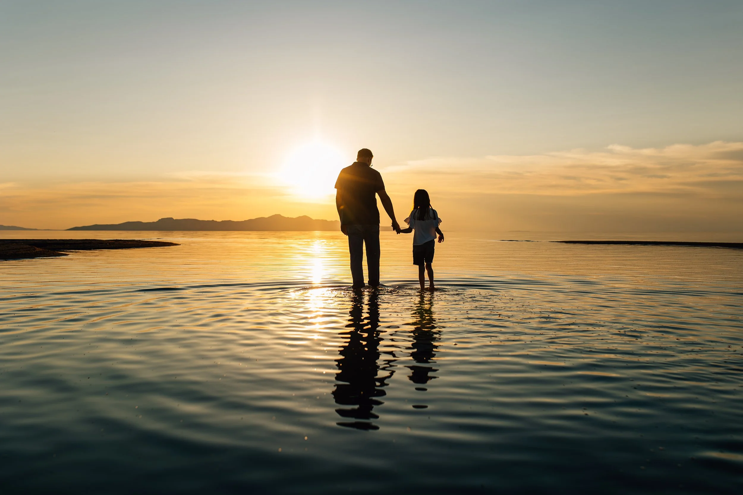 Documentary family photograph of a parent and child holding hands in shallow water at sunset near the Great Salt Lake in Utah