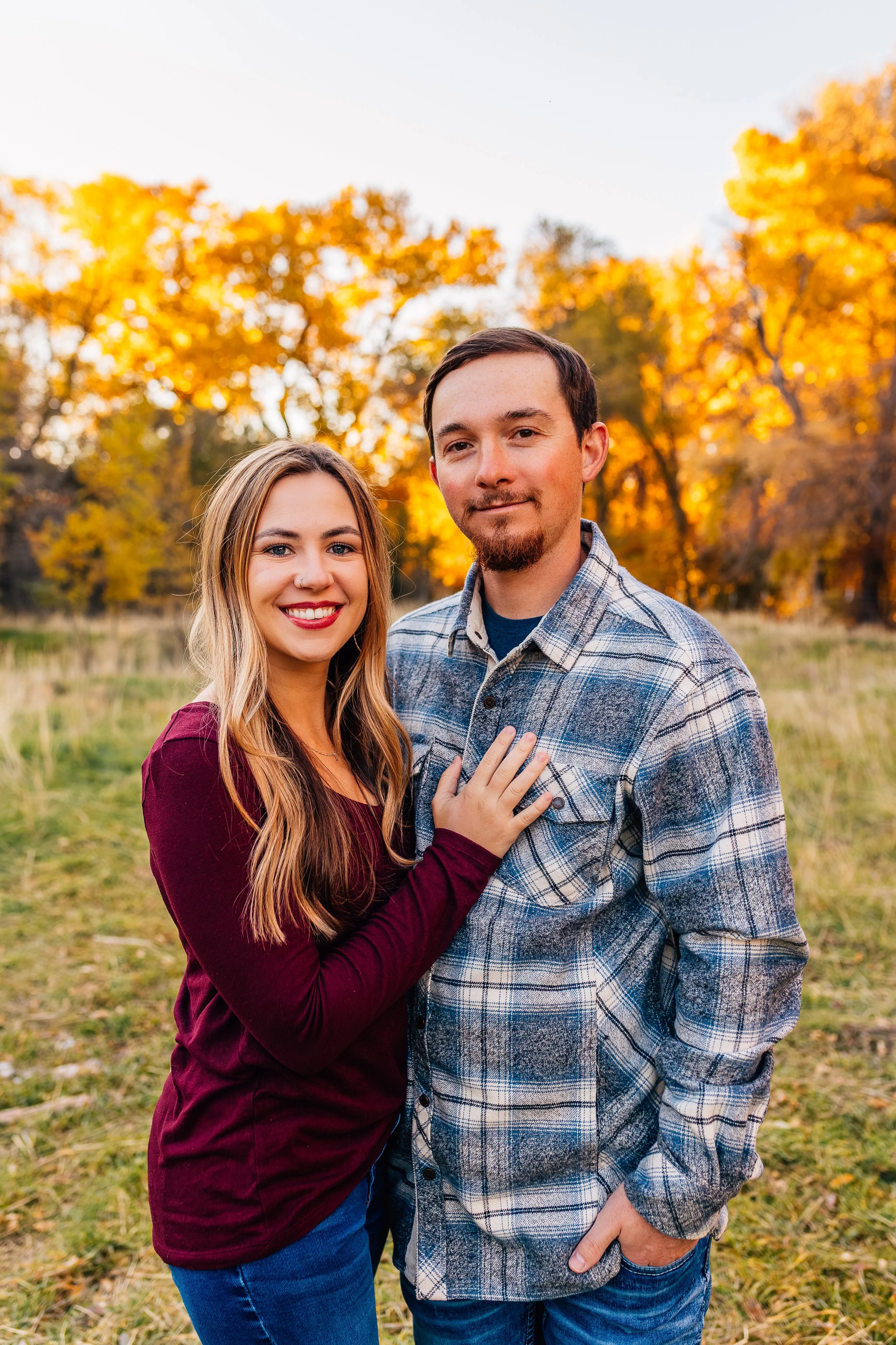 A young couple standing together outdoors during fall, with colorful autumn trees in the background. The woman has long blonde hair and is wearing a maroon long-sleeve top, while the man has short dark hair, a beard, and is wearing a blue plaid shirt